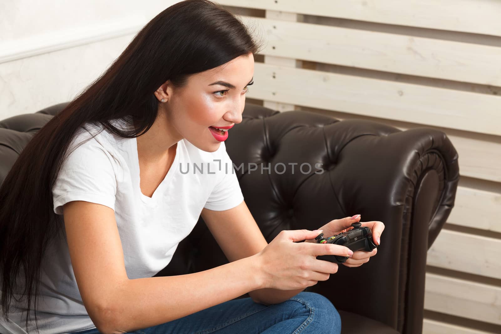 Woman gamer sitting on brown leather sofa and playing a video ga by Nobilior