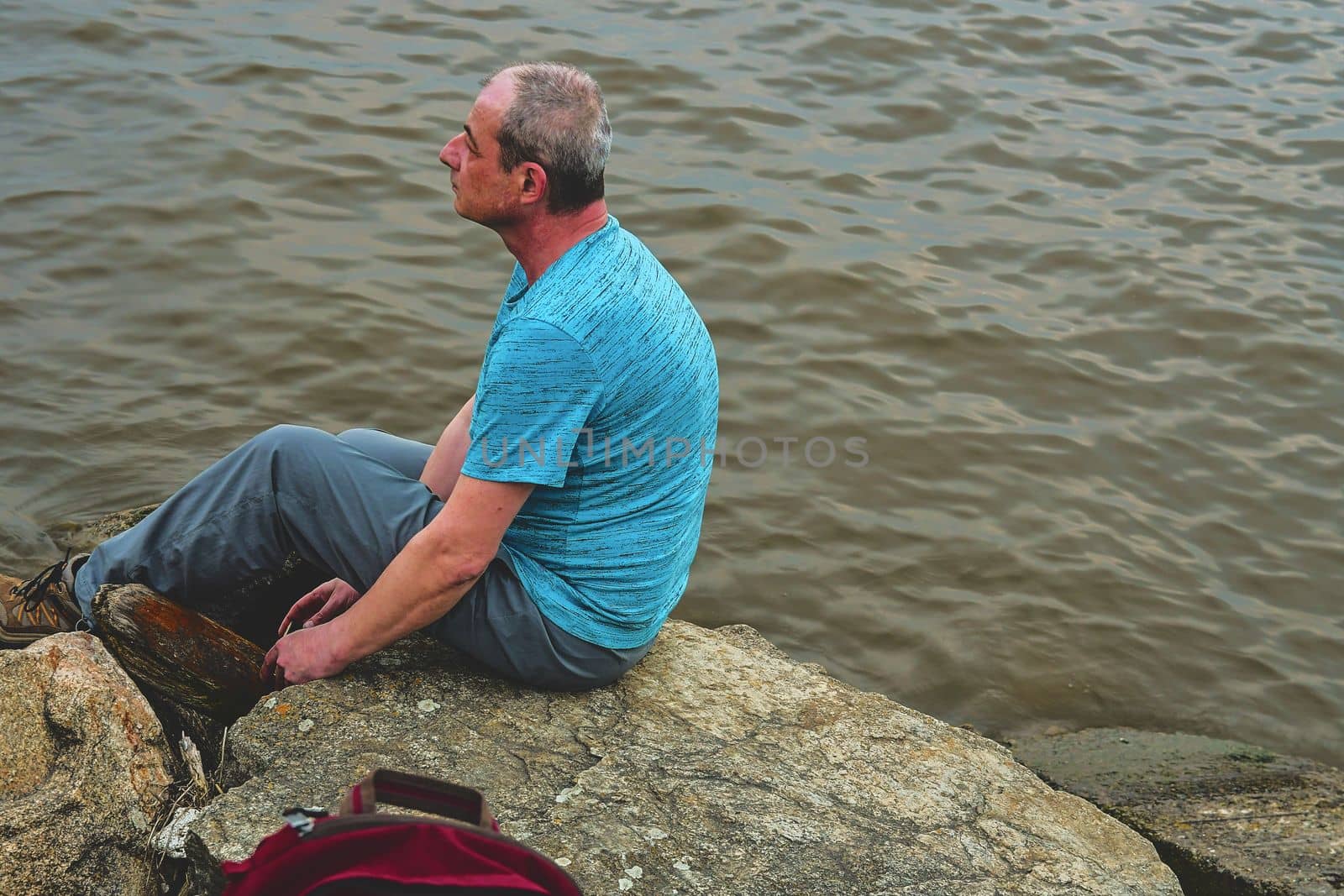 Midle aged man sitting on the shore of the lake. Solitude mature man sitting on the bank. Concept of loneliness. 