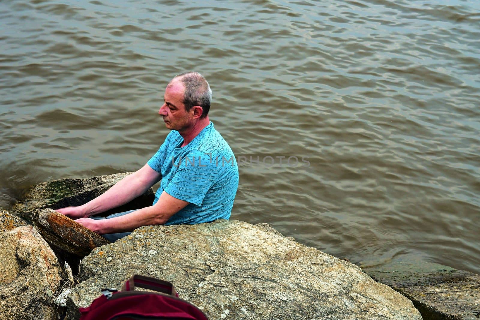 Midle aged man sitting on the shore of the lake. Solitude mature man sitting on the bank. Concept of loneliness by roman_nerud