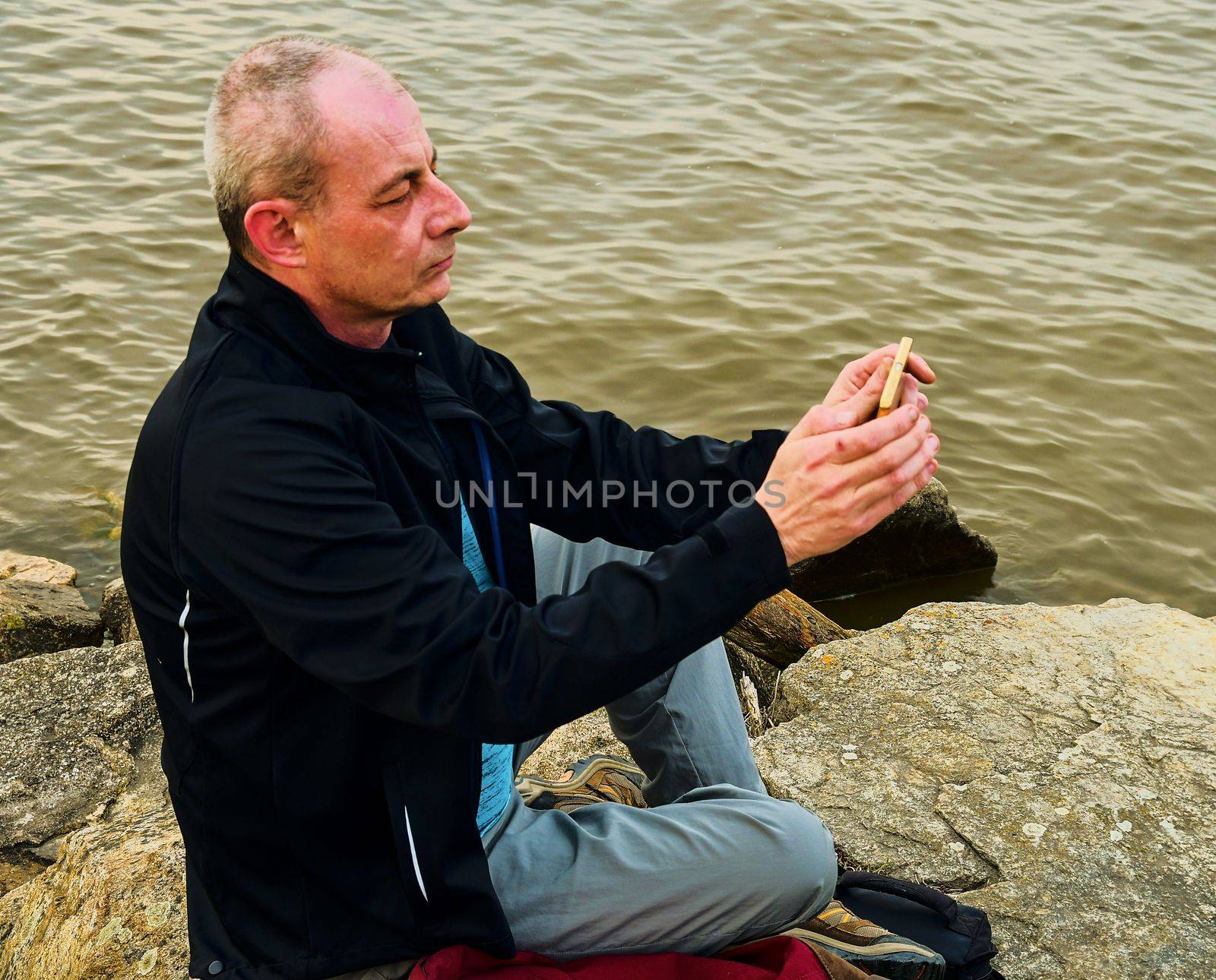 Midle aged man sitting on the shore of the lake. Solitude mature man taking selfie picture  on the bank. Concept of loneliness. 