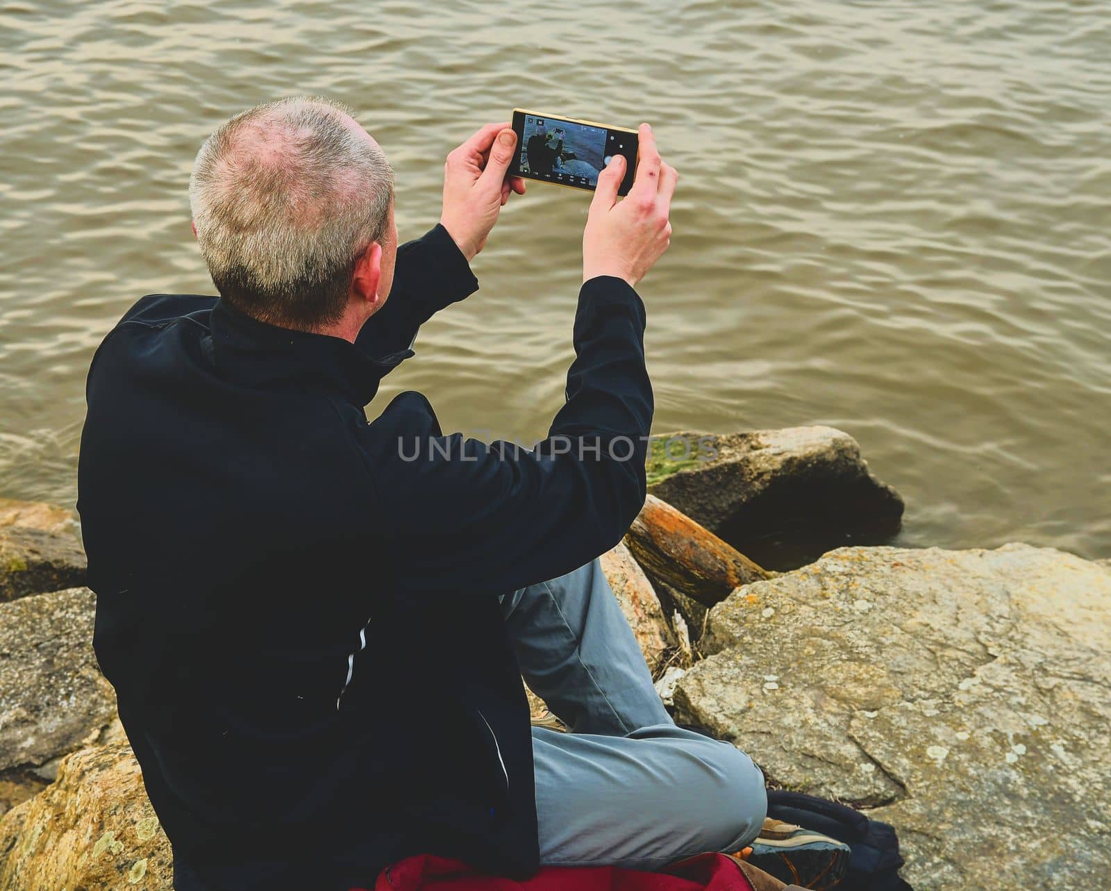 Midle aged man sitting on the shore of the lake. Solitude mature man taking selfie picture  on the bank. Concept of loneliness. 