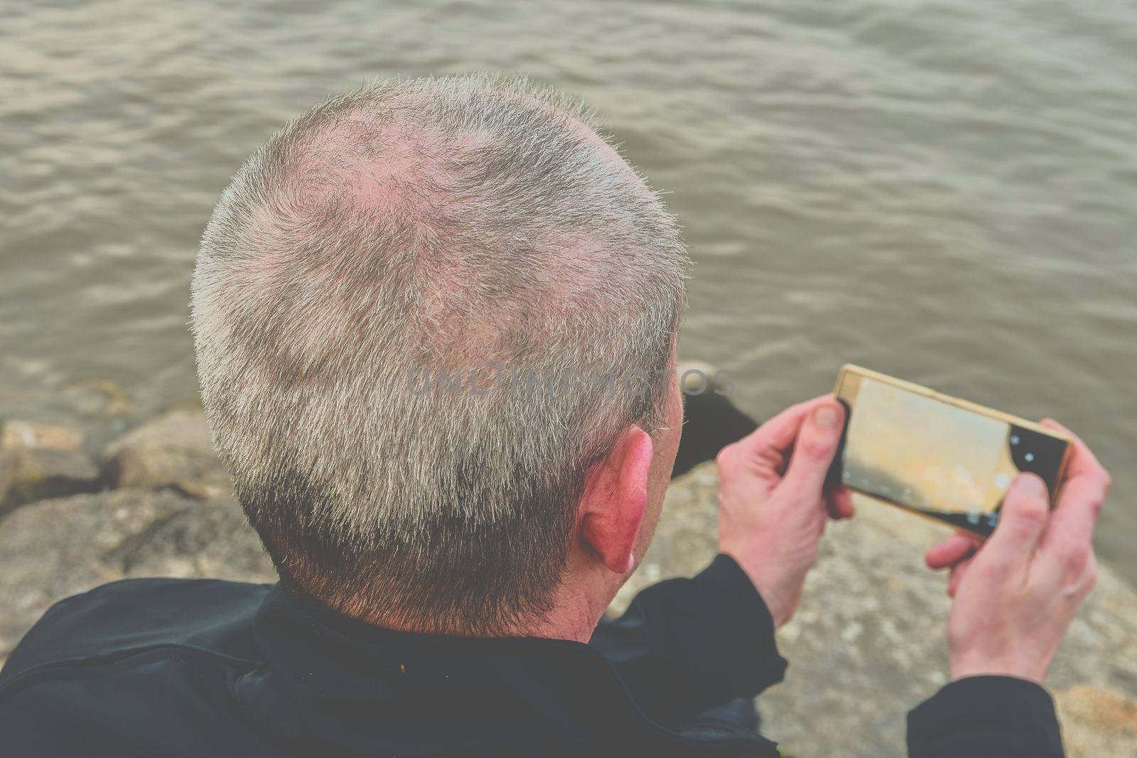 Midle aged man sitting on the shore of the lake. Solitude mature man taking selfie picture  on the bank. Concept of loneliness. 