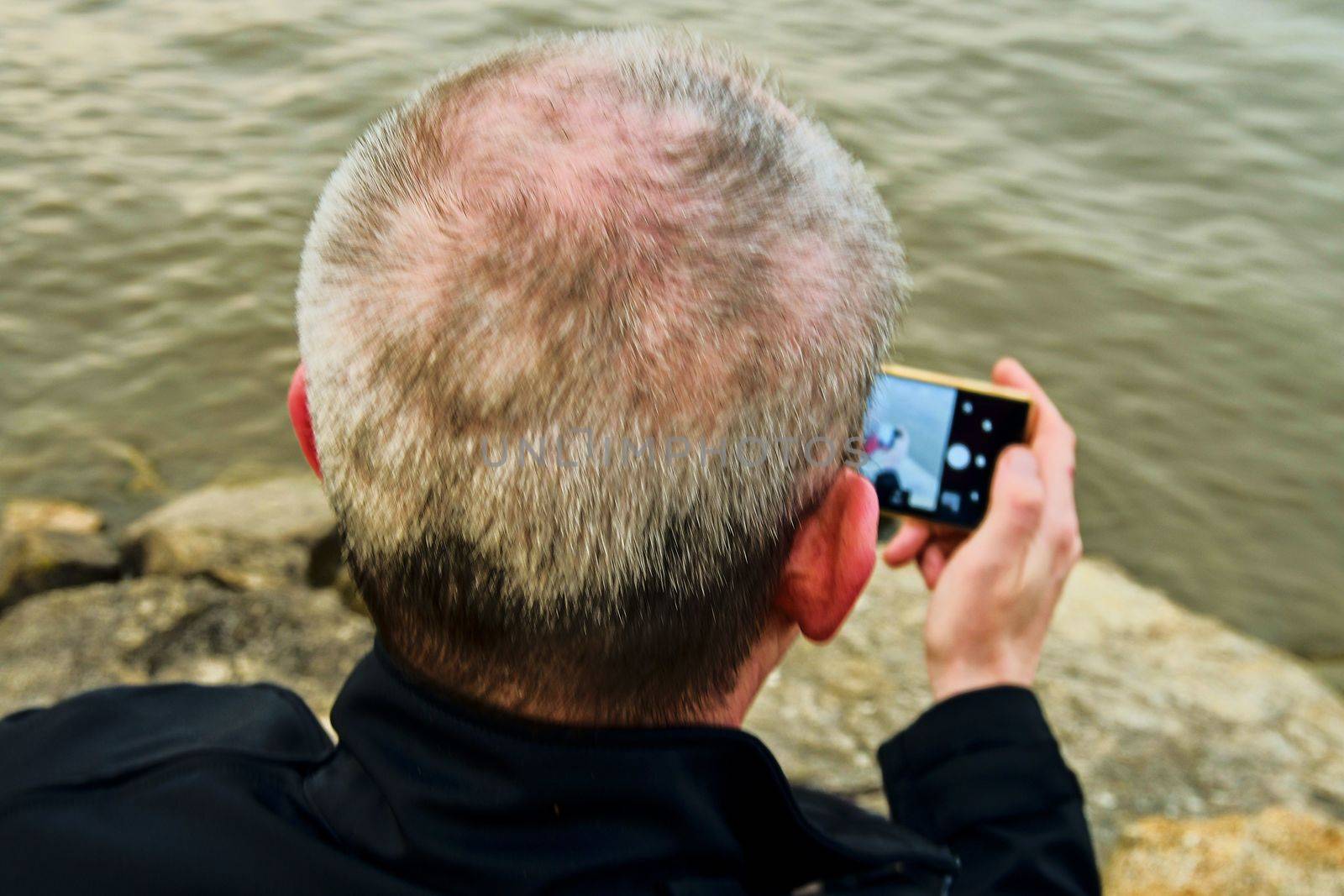 Midle aged man sitting on the shore of the lake. Solitude mature man taking selfie picture  on the bank. Concept of loneliness. 