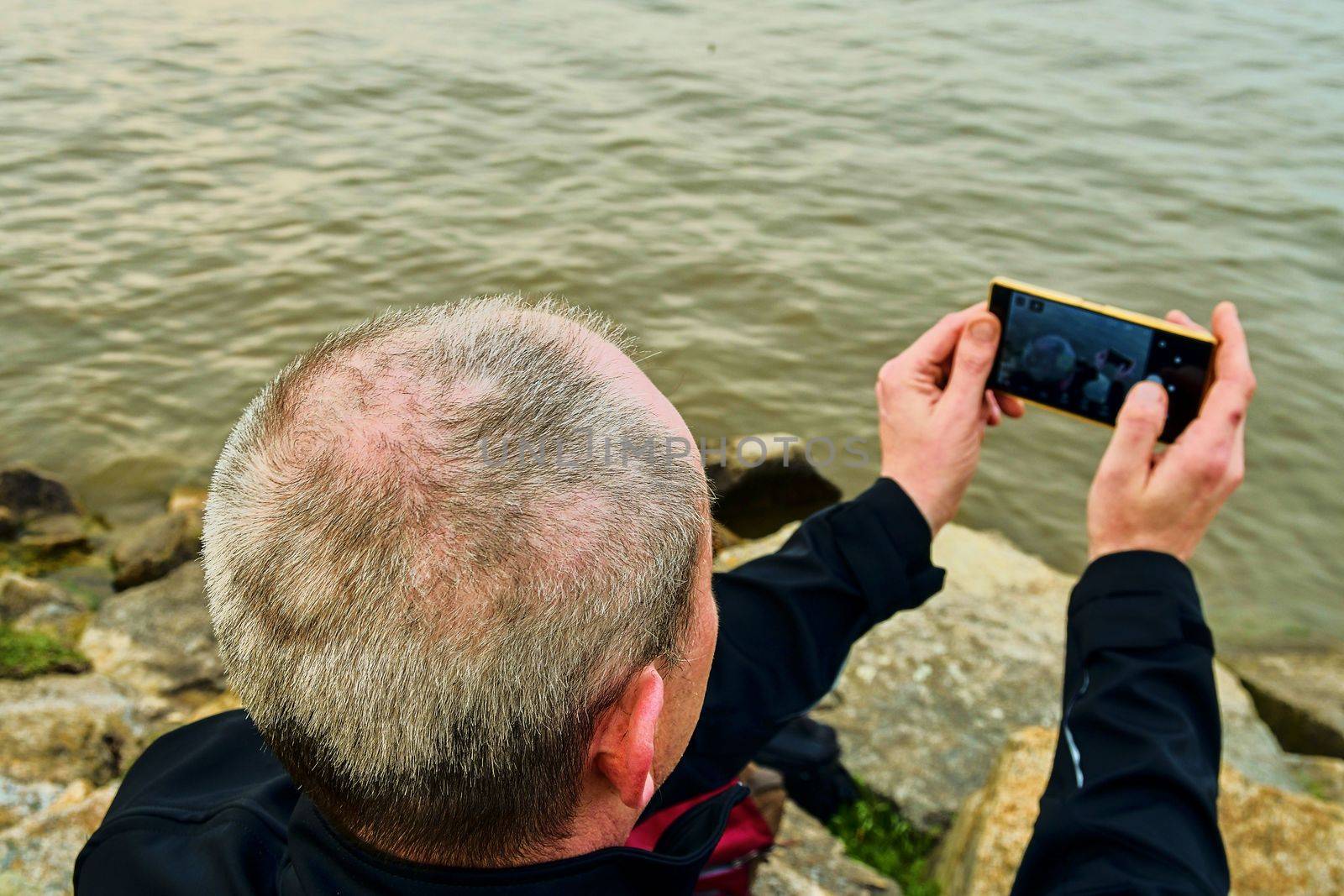 Midle aged man sitting on the shore of the lake. Solitude mature man taking selfie picture  on the bank. Concept of loneliness by roman_nerud