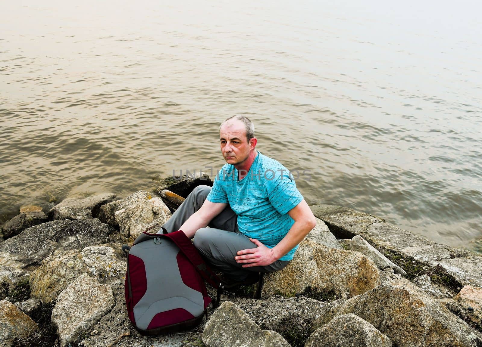 Midle aged man sitting on the shore of the lake. Solitude mature man sitting on the bank. Concept of loneliness. 