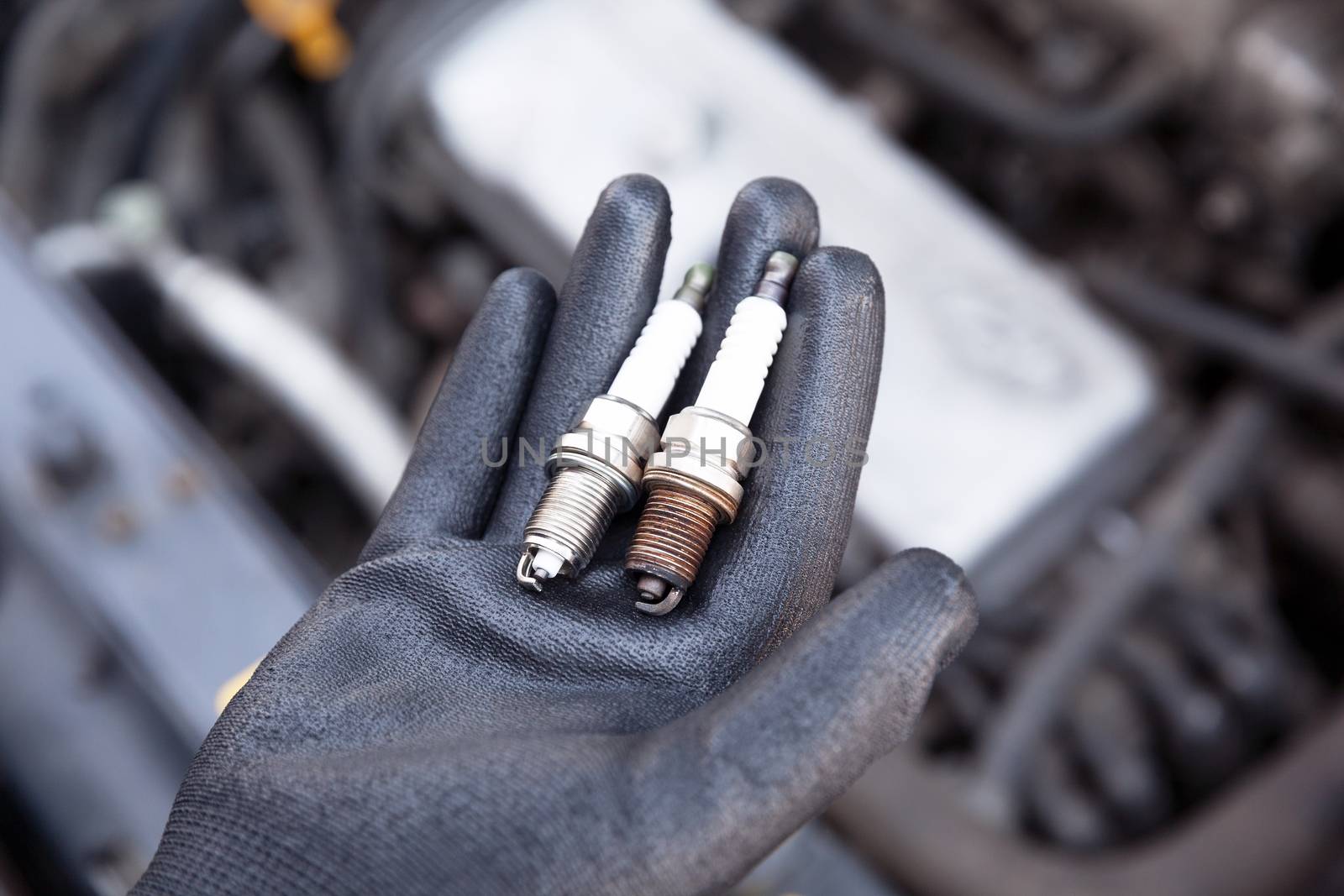 Auto mechanic wearing protective work glove holds old and new spark plugs over a car engine