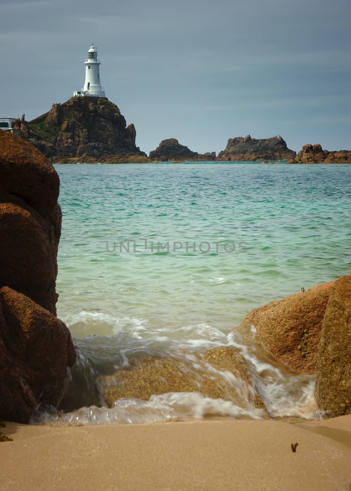 Calm sea on a summer day at Corbiere Lighthouse, Jersey, UK. A small wave is splashing onto the beach. The lighthouse is in the ackground.