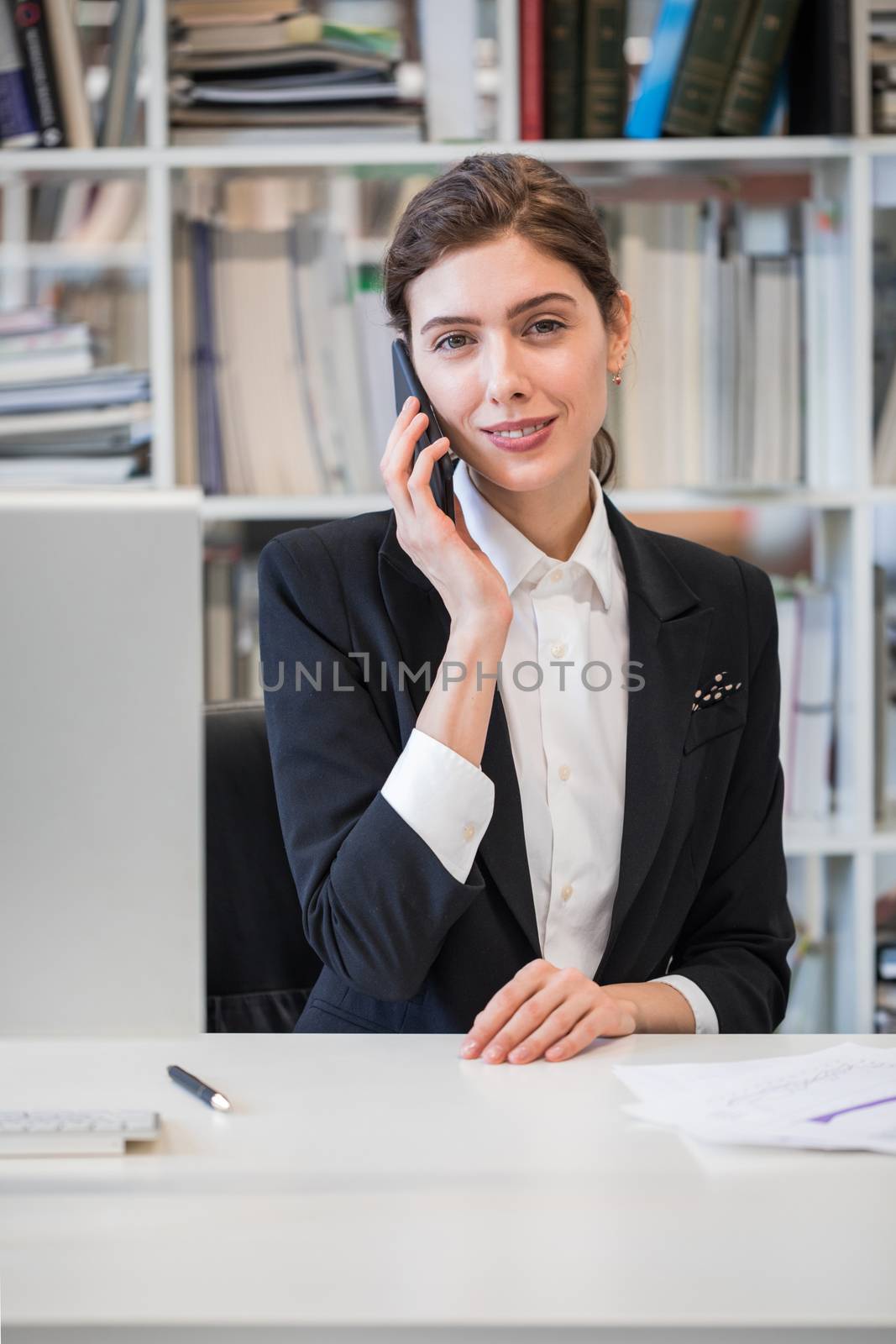 Beautiful mid age business woman talking on phone and looking at camera sitting at her working place in office