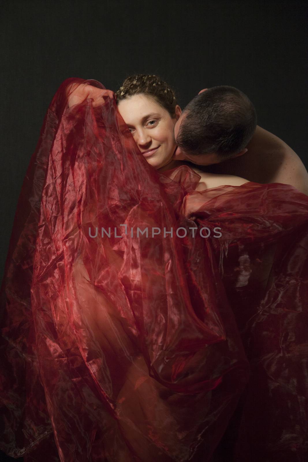 Happy pregnant woman covered with red veil is being kissed by a male.