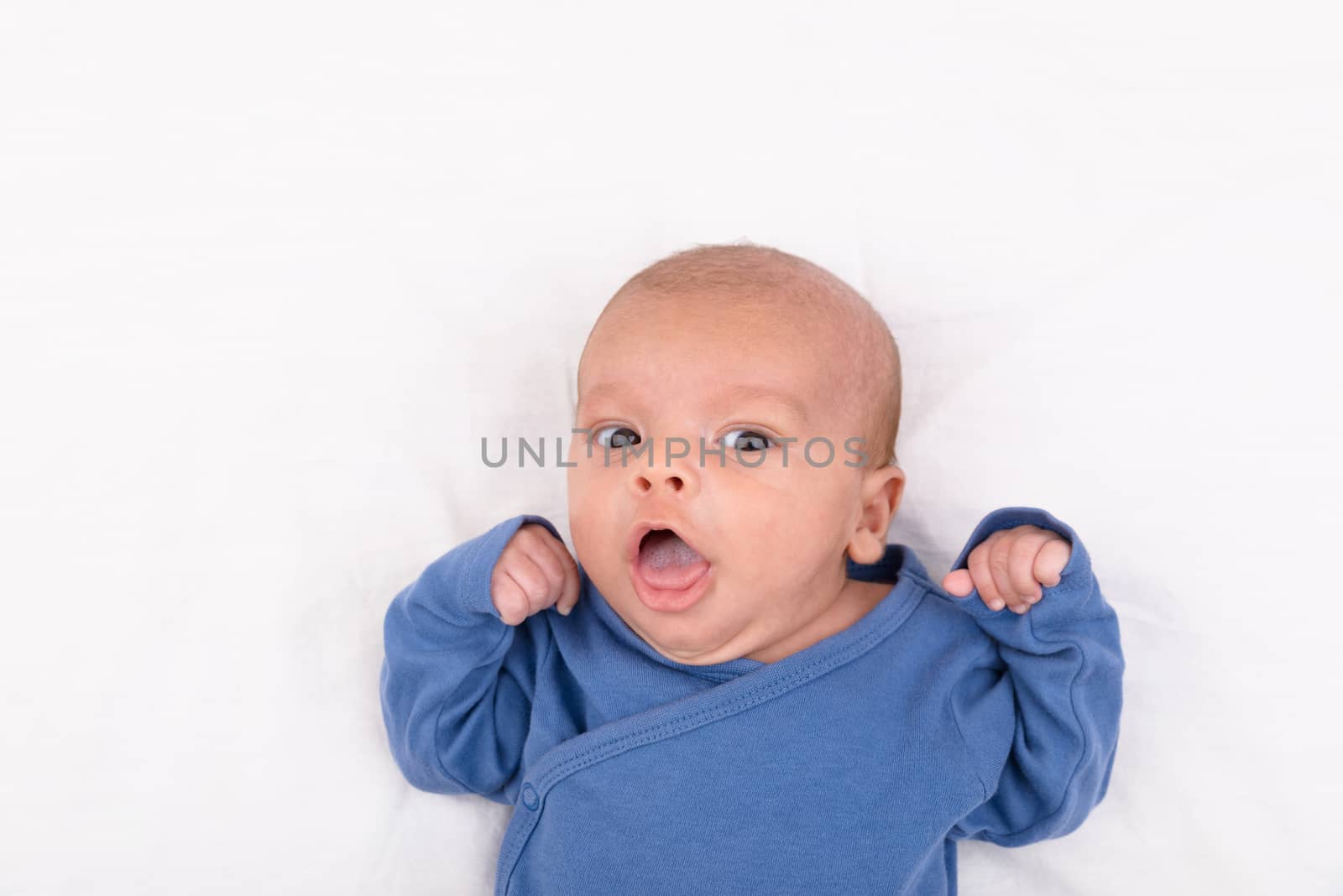 Cute eurasian newborn baby boy wearing a blue infant bodysuit on white sheet and looking at the camera, with copy space