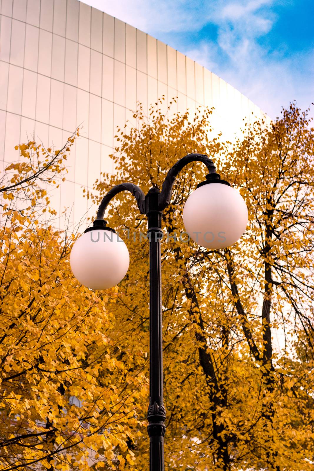 Street lamp in the park with yellow autumn trees and a large building on the background