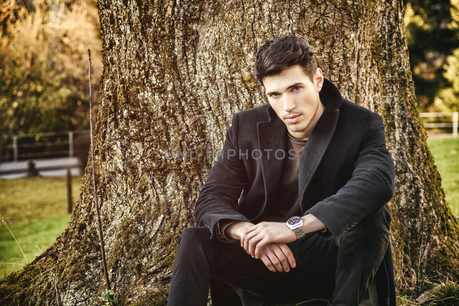 Handsome young man leaning against tree, looking at camera, in a sunny day wearing a black coat