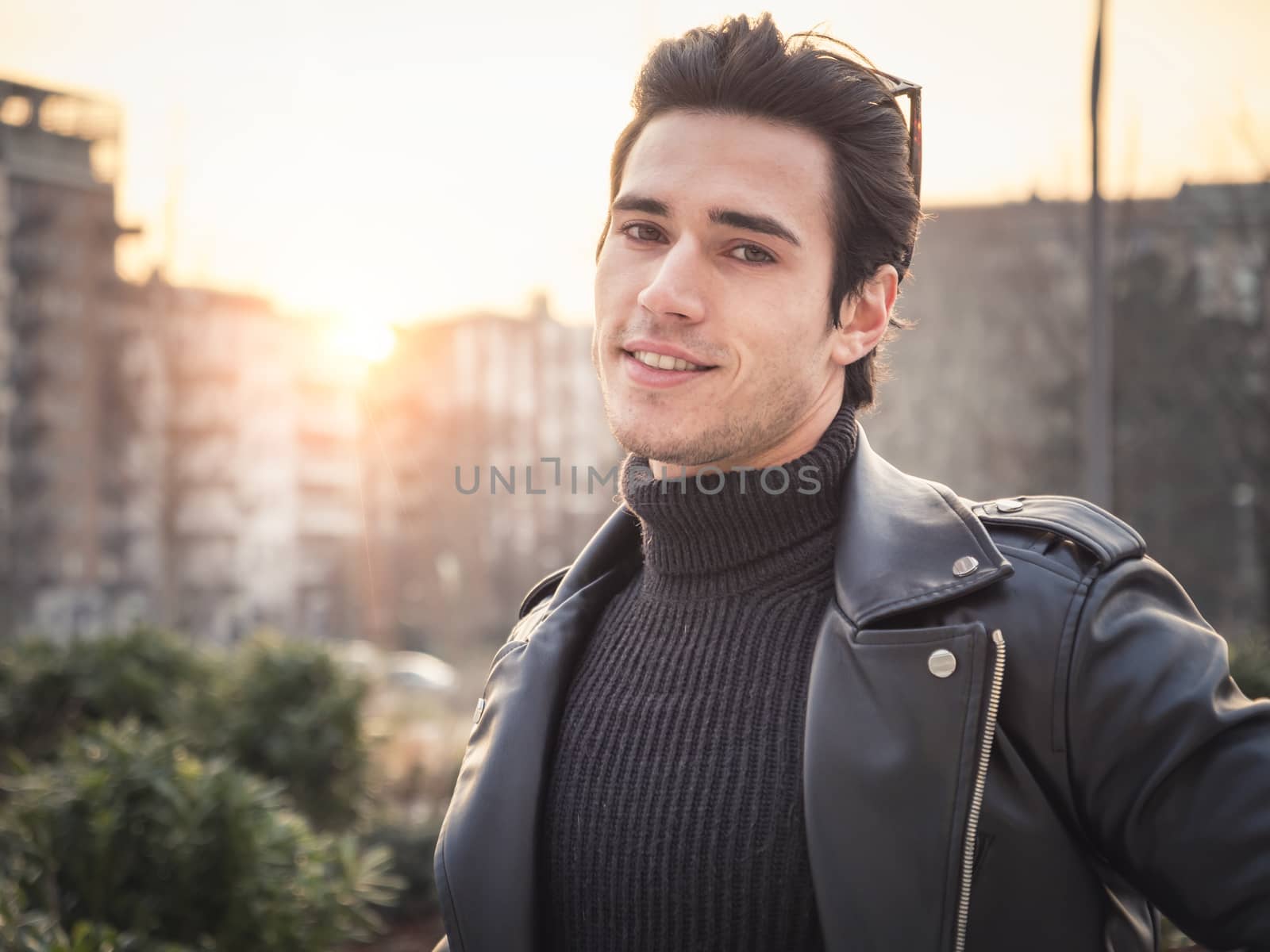 One handsome young man in urban setting in modern city, standing, wearing black leather jacket and jeans, looking at camera, at sunset