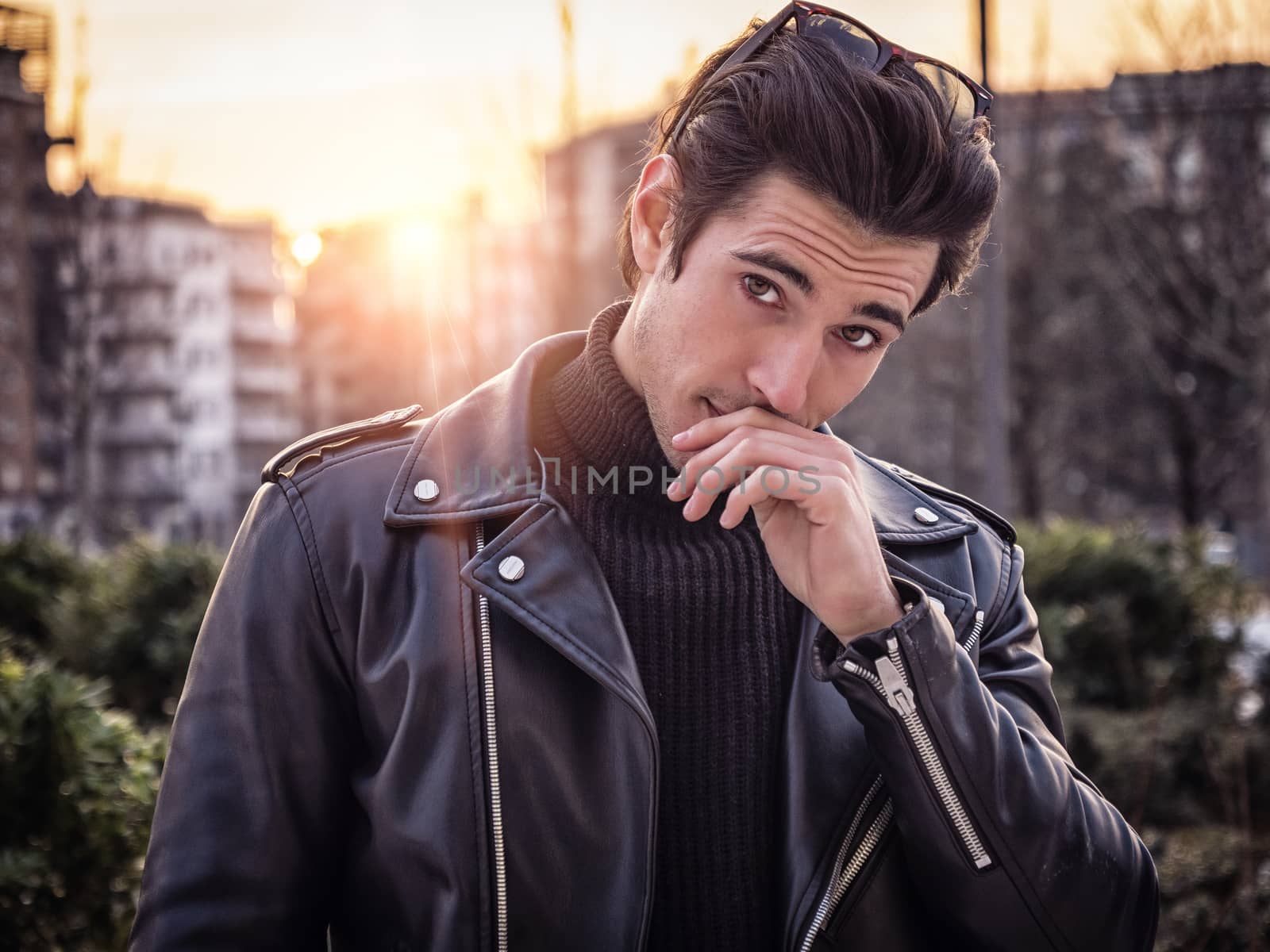 One handsome young man in urban setting in modern city, standing, wearing black leather jacket and jeans, looking at camera, at sunset