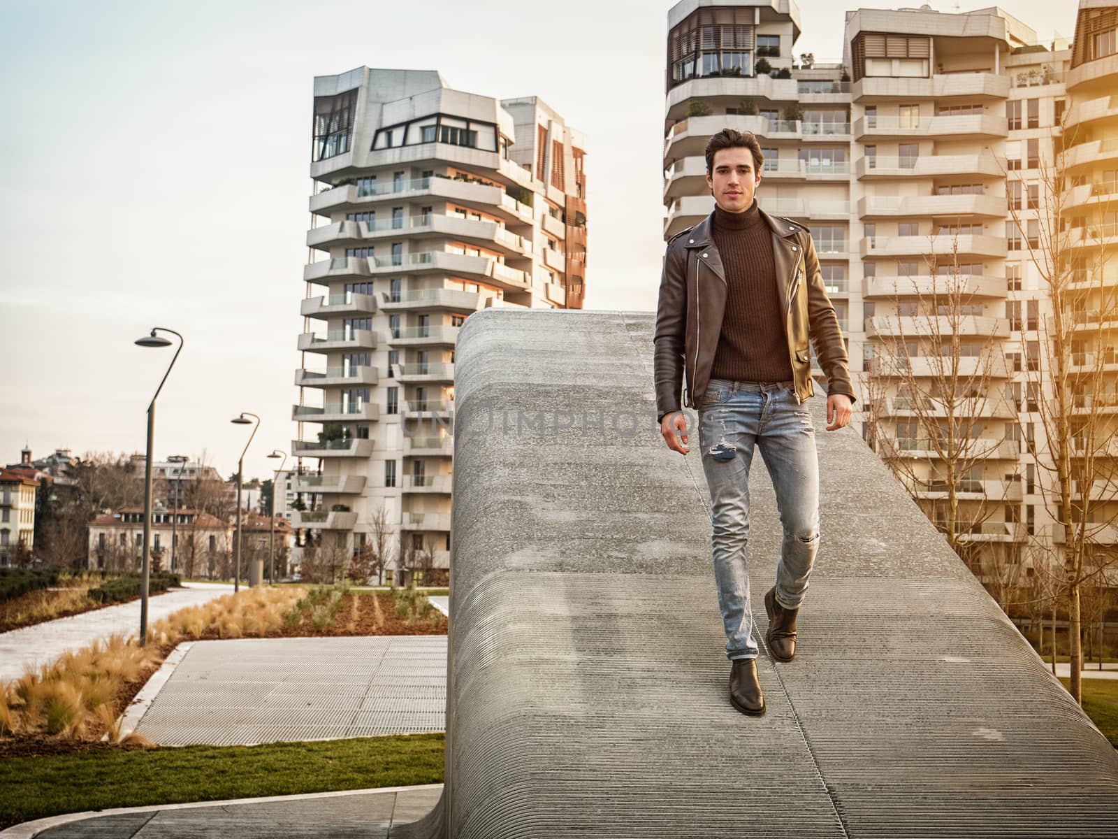 One handsome young man in urban setting in modern city, standing, wearing black leather jacket and jeans, looking at camera