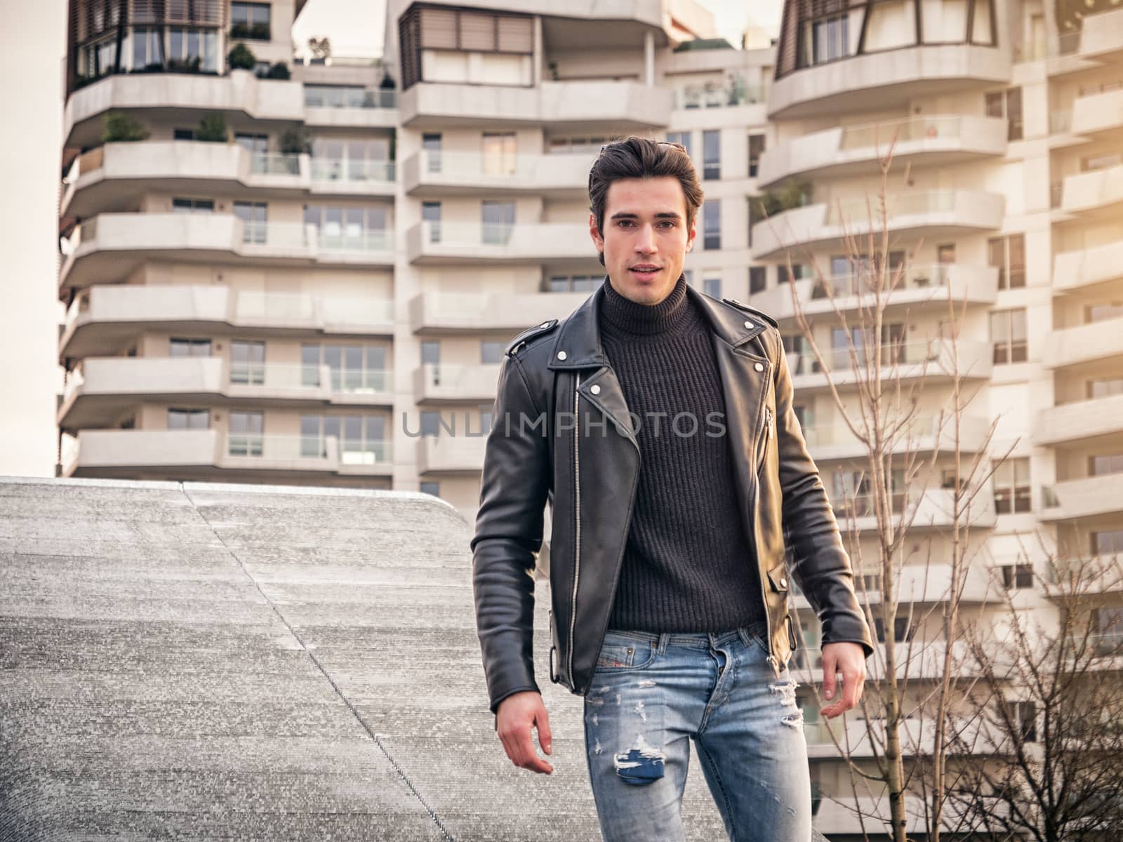 One handsome young man in urban setting in modern city, standing, wearing black leather jacket and jeans, looking at camera