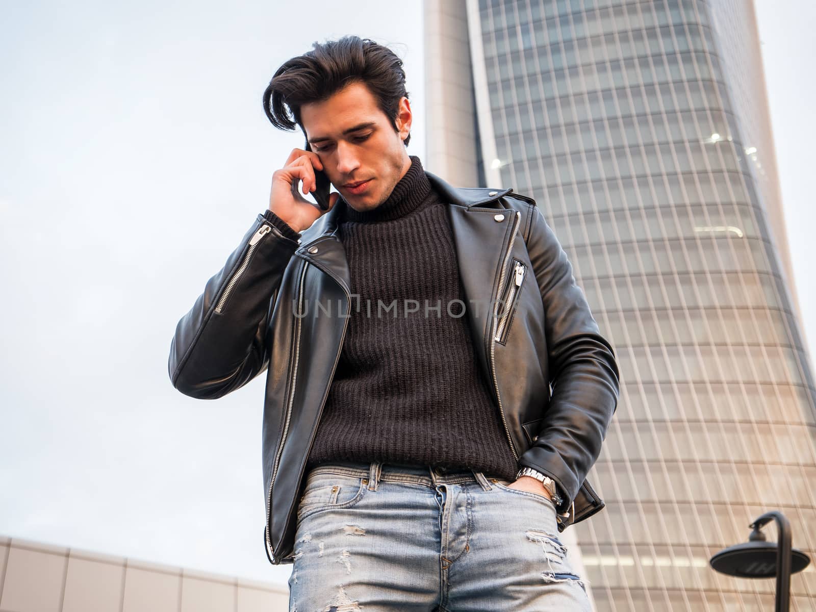 One handsome young man in urban setting in modern city, calling with cellphone, standing, wearing black leather jacket and jeans
