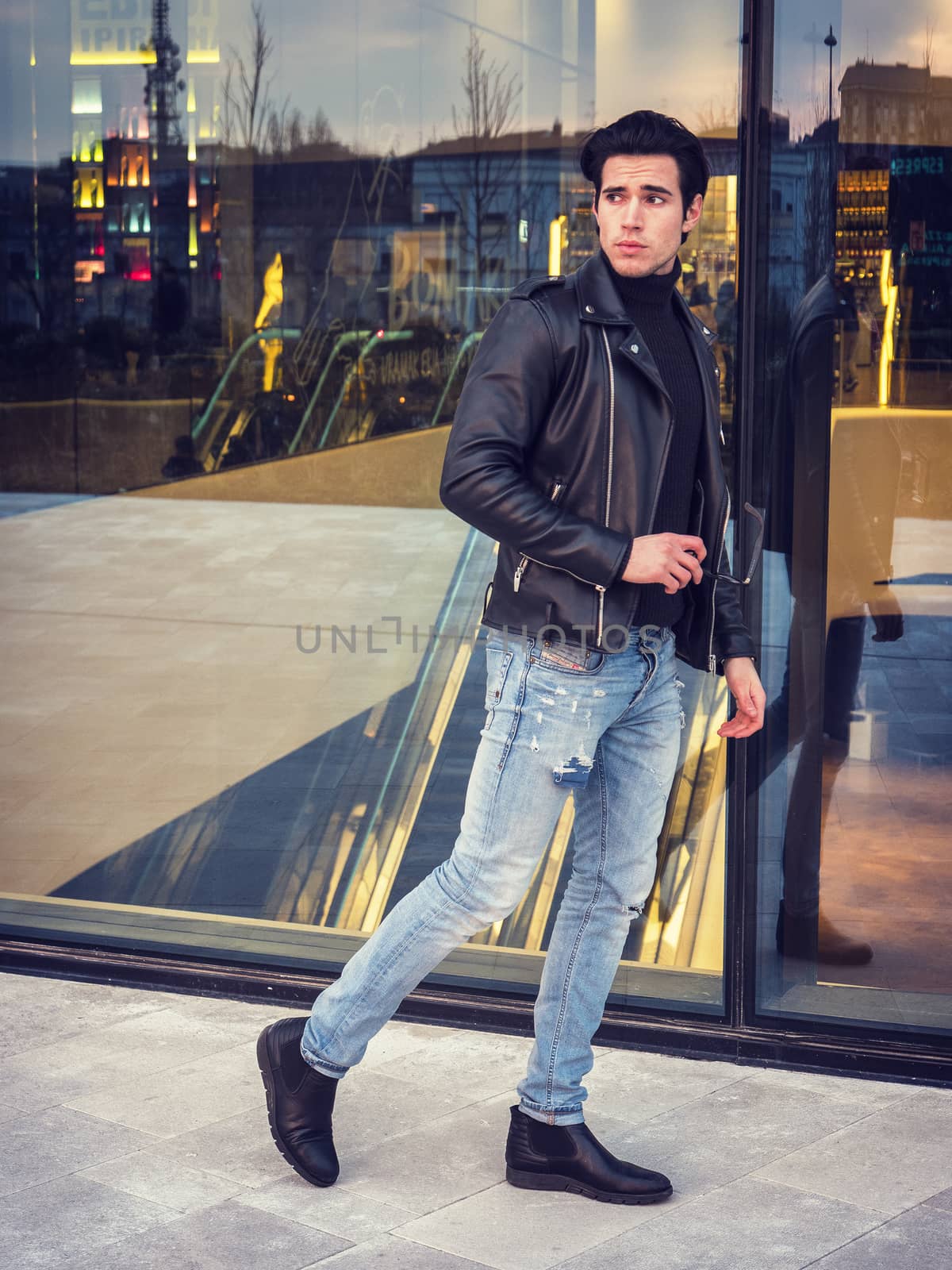 One handsome young man in urban setting in modern city, walking, wearing black leather jacket and jeans