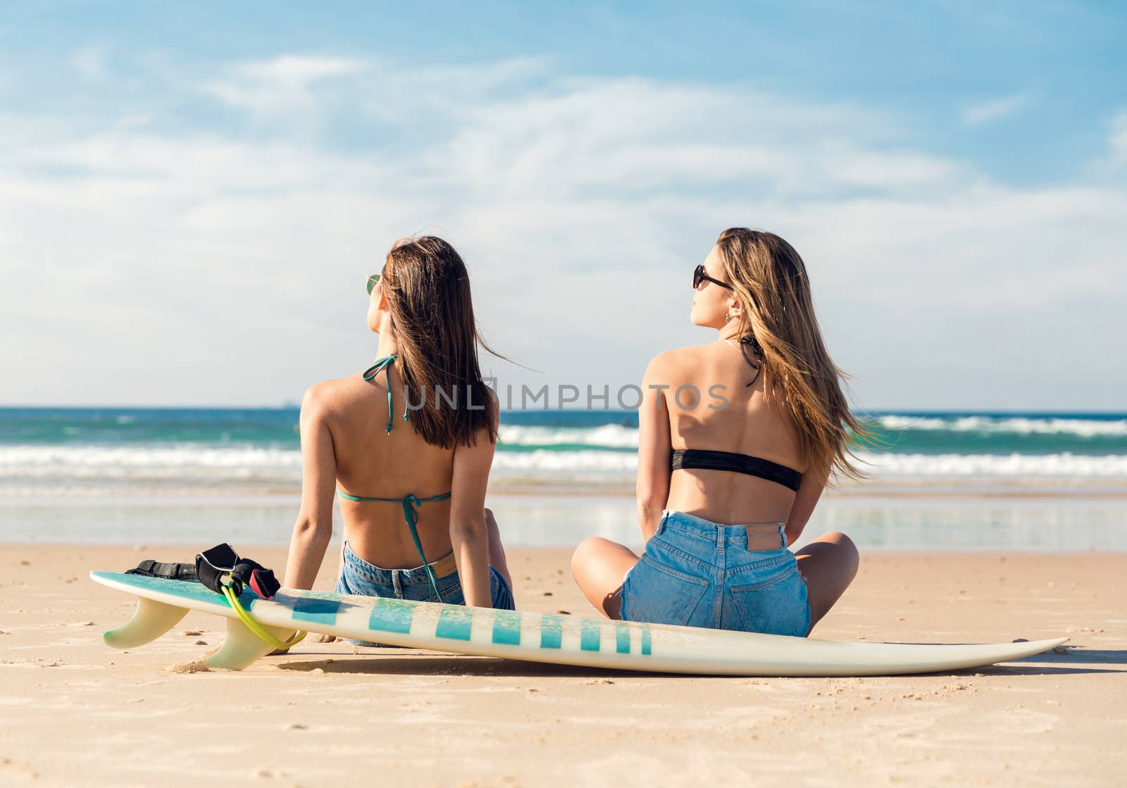 Two beautiful female friends at the beach sitting on the sand close to her surfboards while looking to the ocean