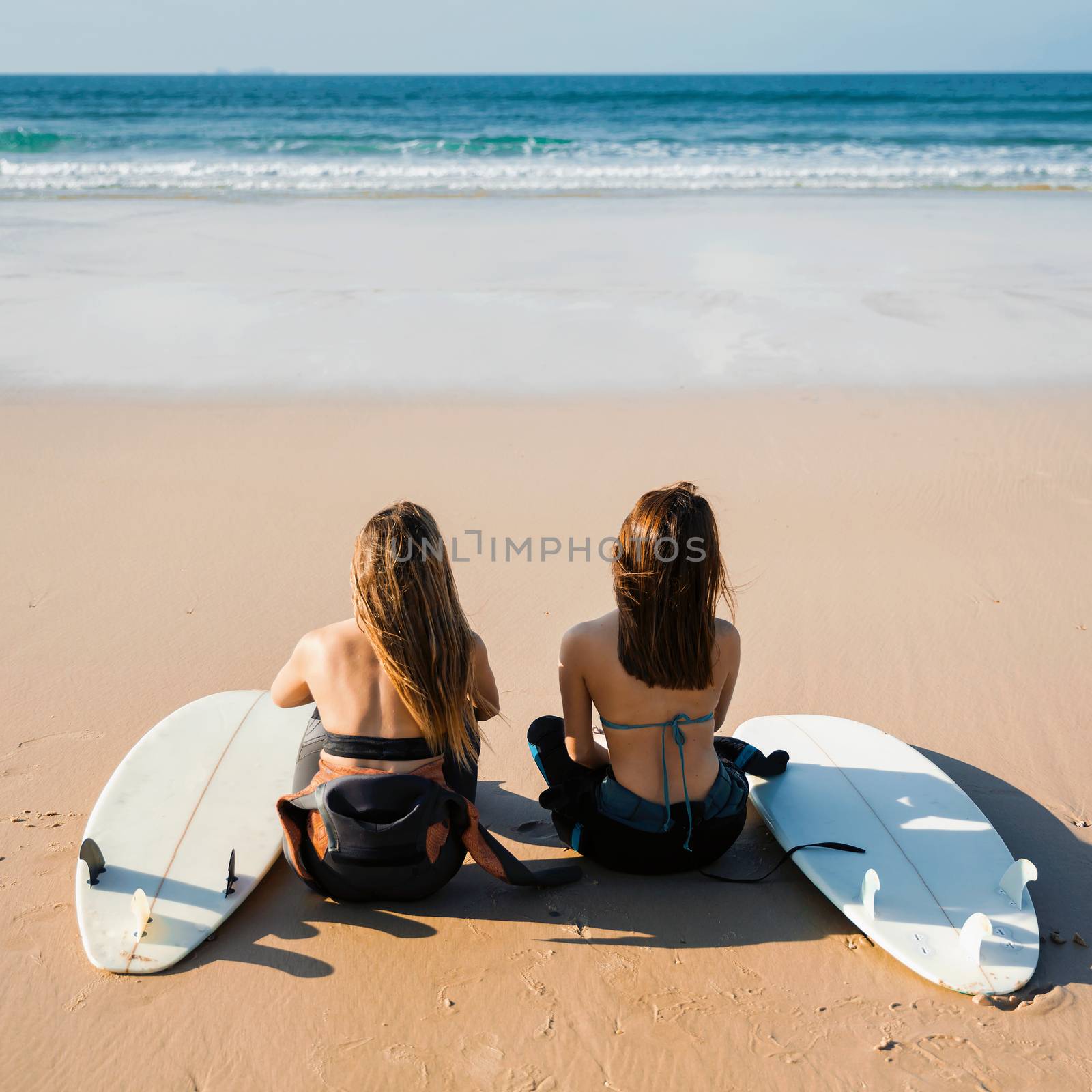 Two beautiful female friends at the beach sitting on the sand with her surfboards while looking to the ocean