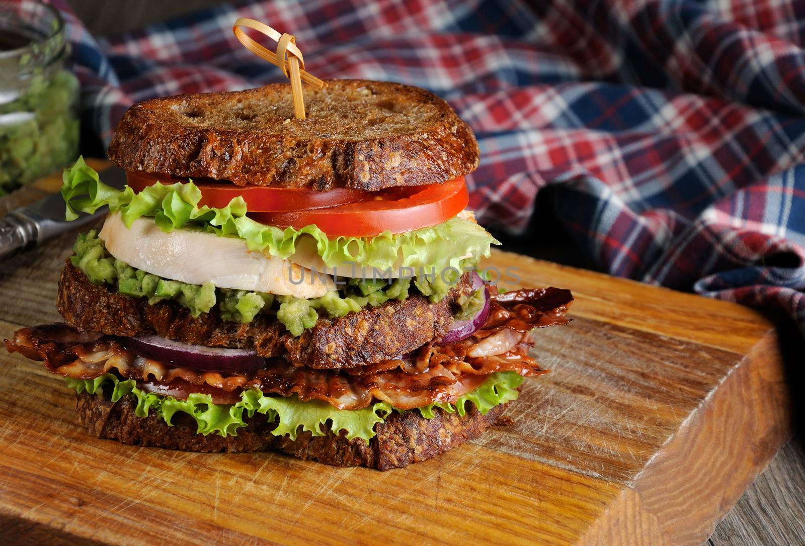 A double sandwich made of Greek bread with slices of fried bacon, avocado, boiled chicken breast, and tomatoes.