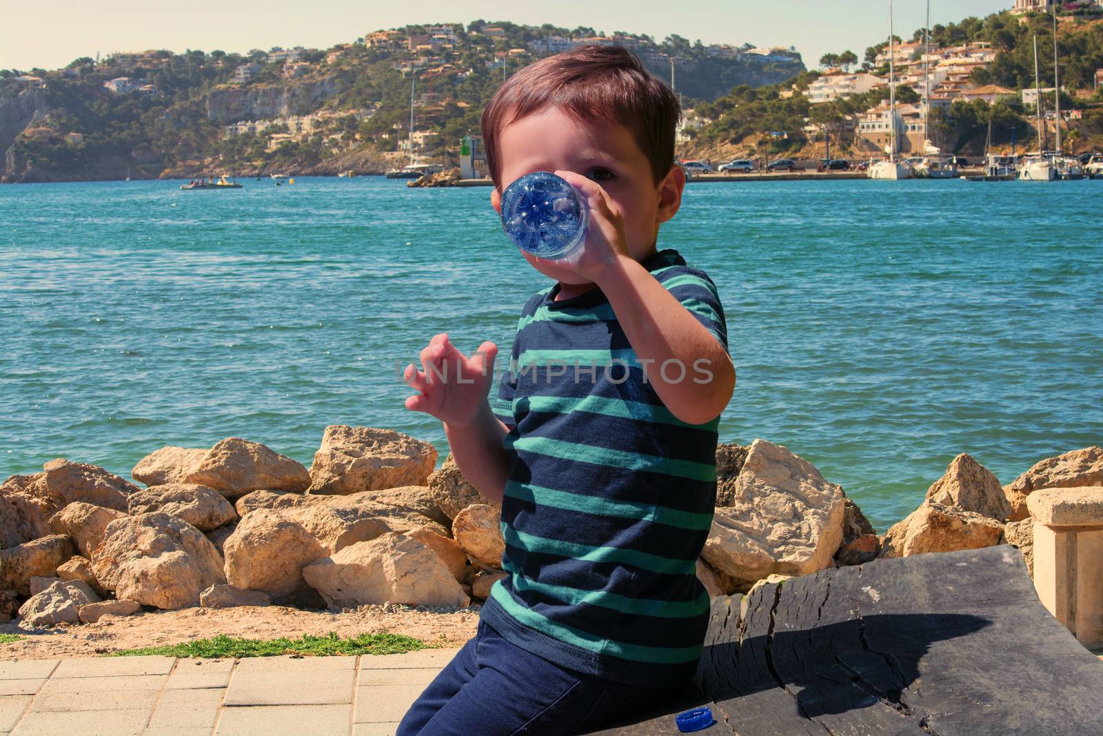 Thirsty little boy sitting on a wooden bench and drinking mineral water. In the background a Mediterranean port.