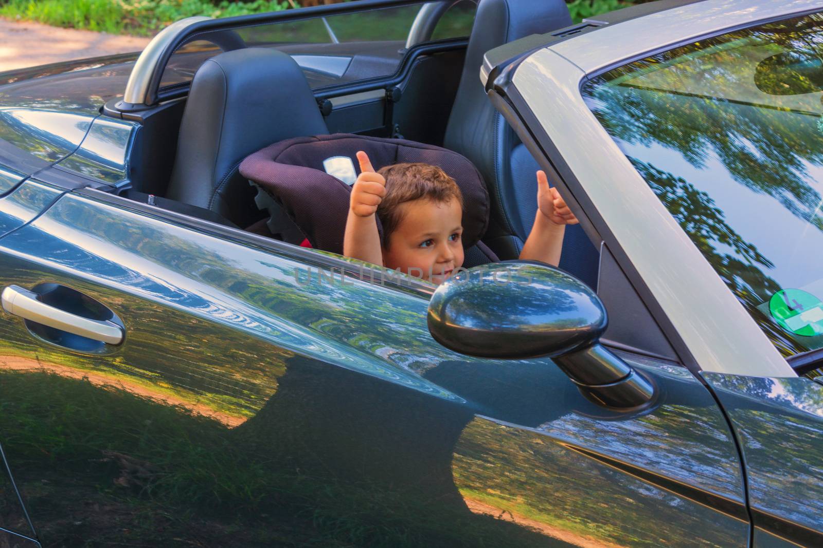 Happy laughing child in a convertible sits in the car child seat and shows the thumbs up.