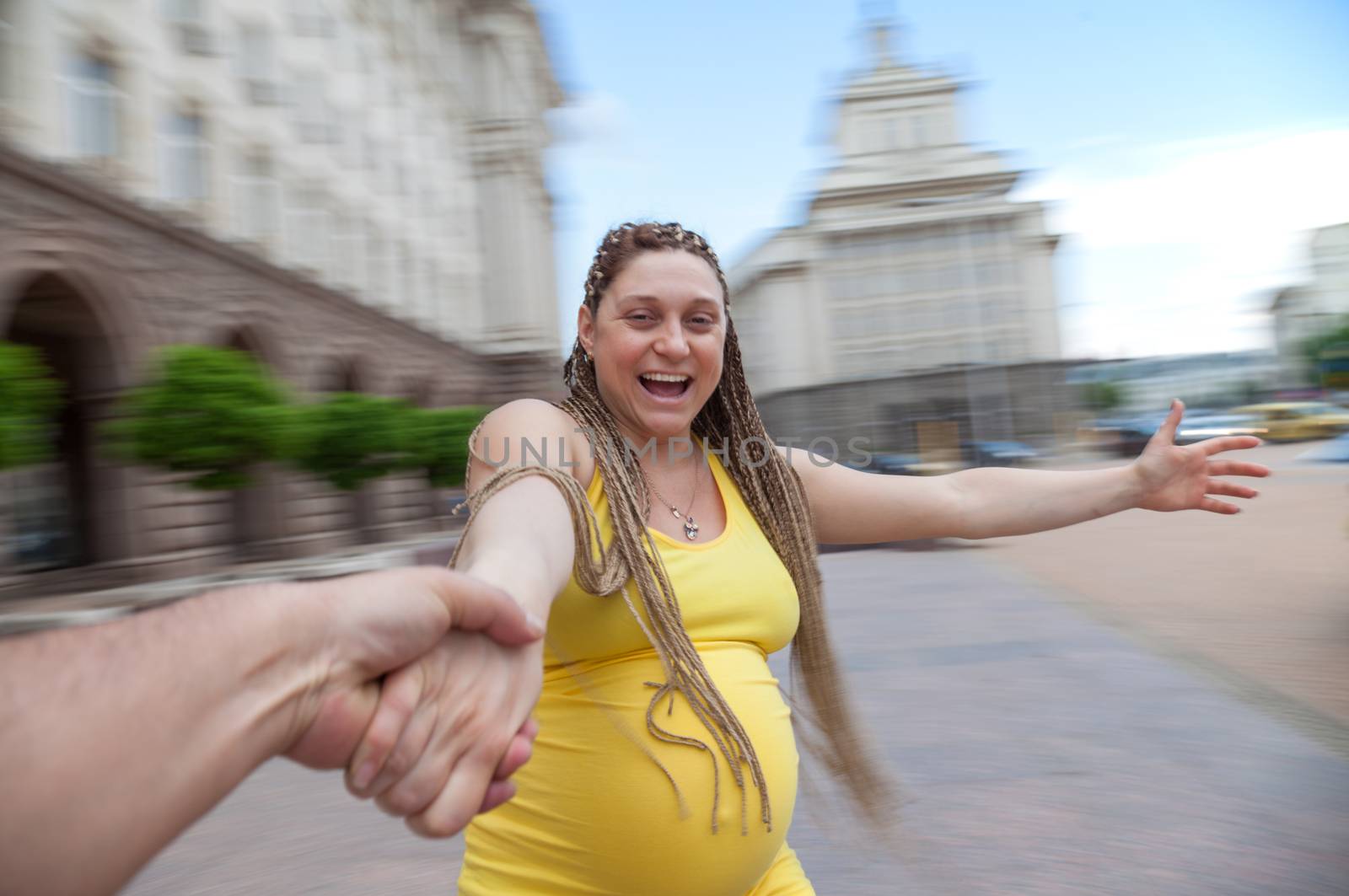 Laughing pregnant woman holding male hand is spinning outdoors.