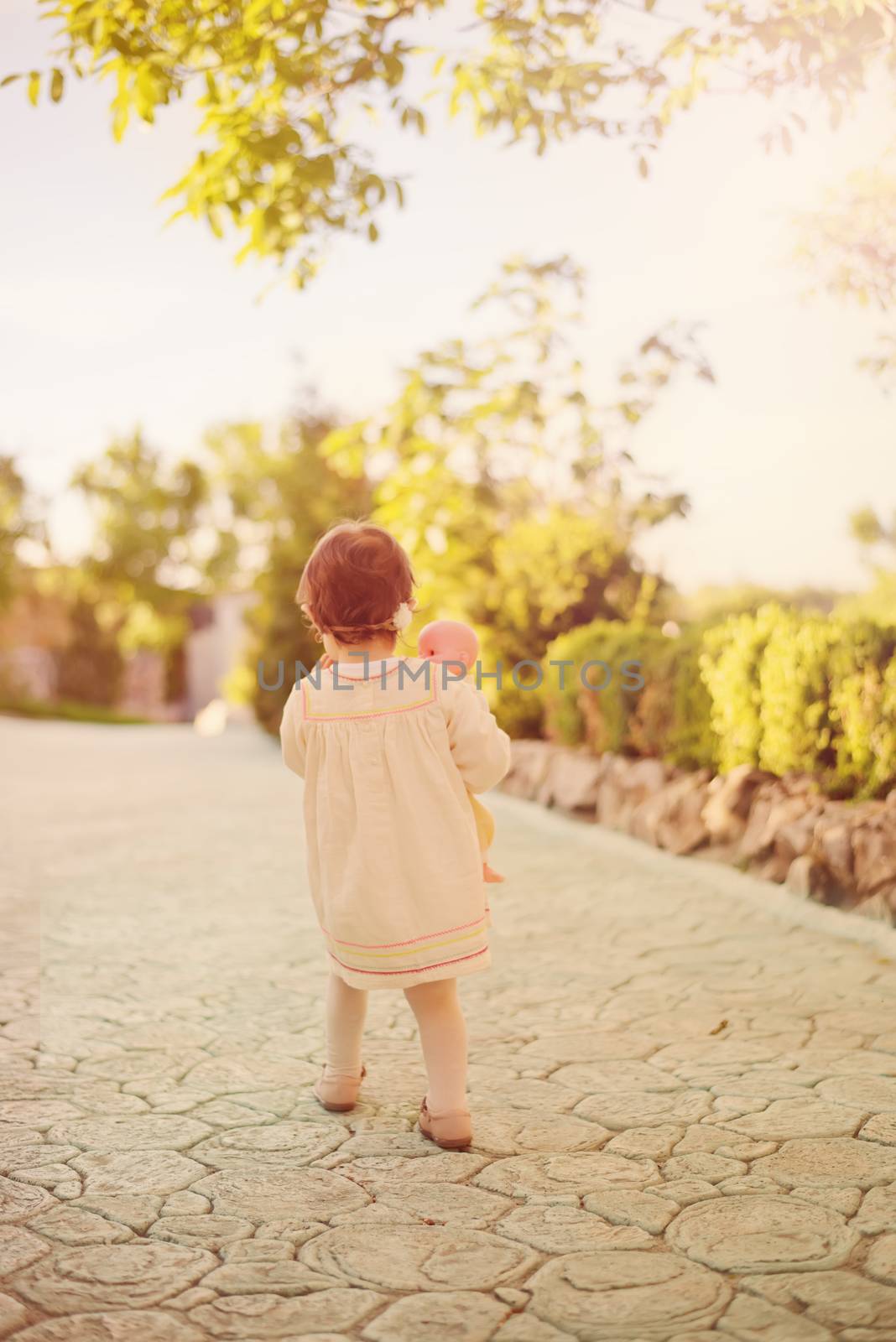 Toddler girl in the back, dressed in a bright dress with a wreath of flowers on his head and holding a doll. Little girl walking down an stone alley in a garden with flowers and trees. Sun, sun haze, glare.