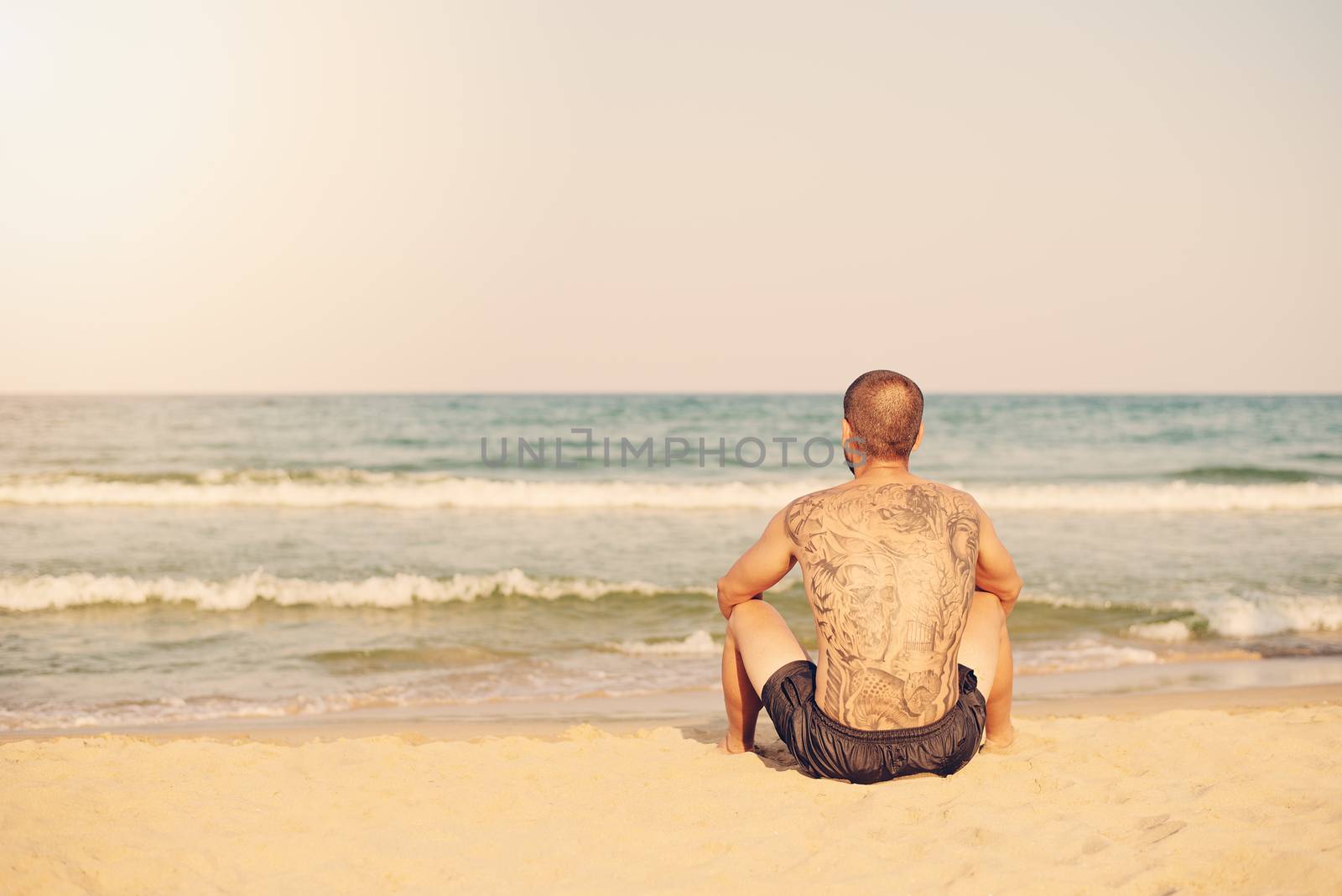 A young man with a large tattoo on his back sitting on the beach overlooking the sea. Sun, sun haze, glare. Copy space on left