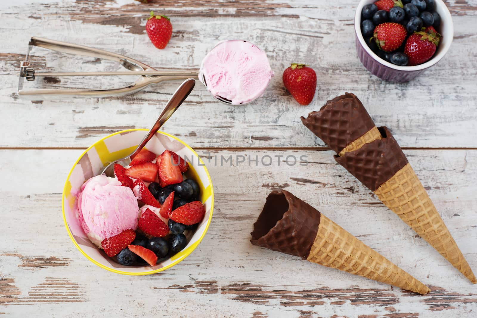 Pink Ice Cream served with berries - strawberries and blueberries in a yellow bowl. Waffle cones with chocolate. Light Rustic Wooden Background