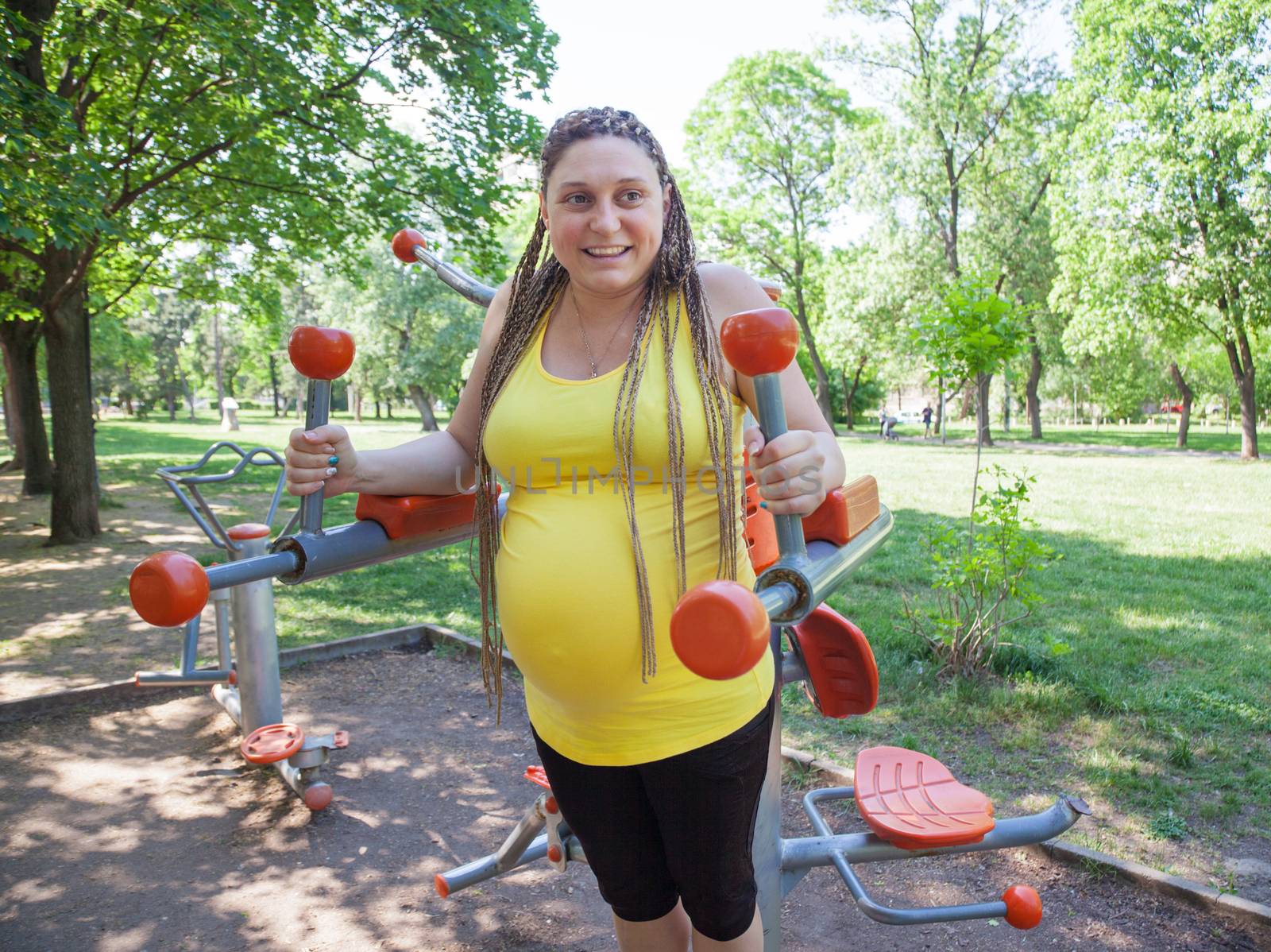 Smiling pregnant woman is doing exercises outdoors on fitness machines in a park.