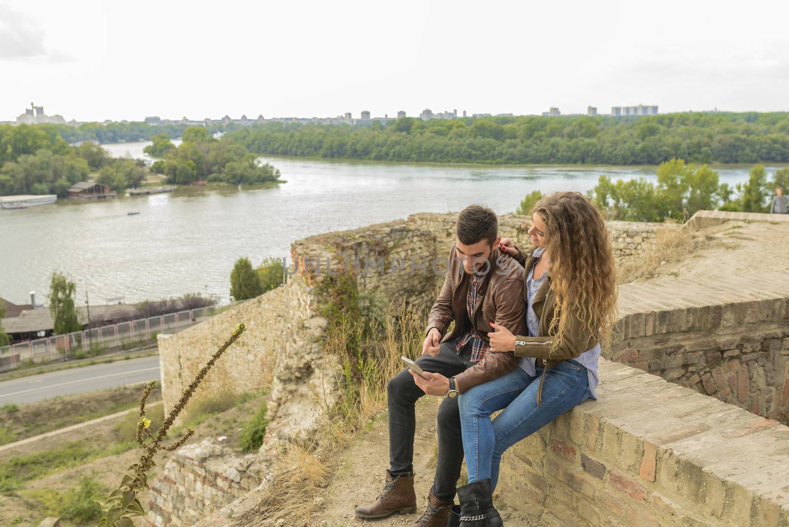 Attractive long haired woman and her boyfriend sitting on the wall and using their smart-phone