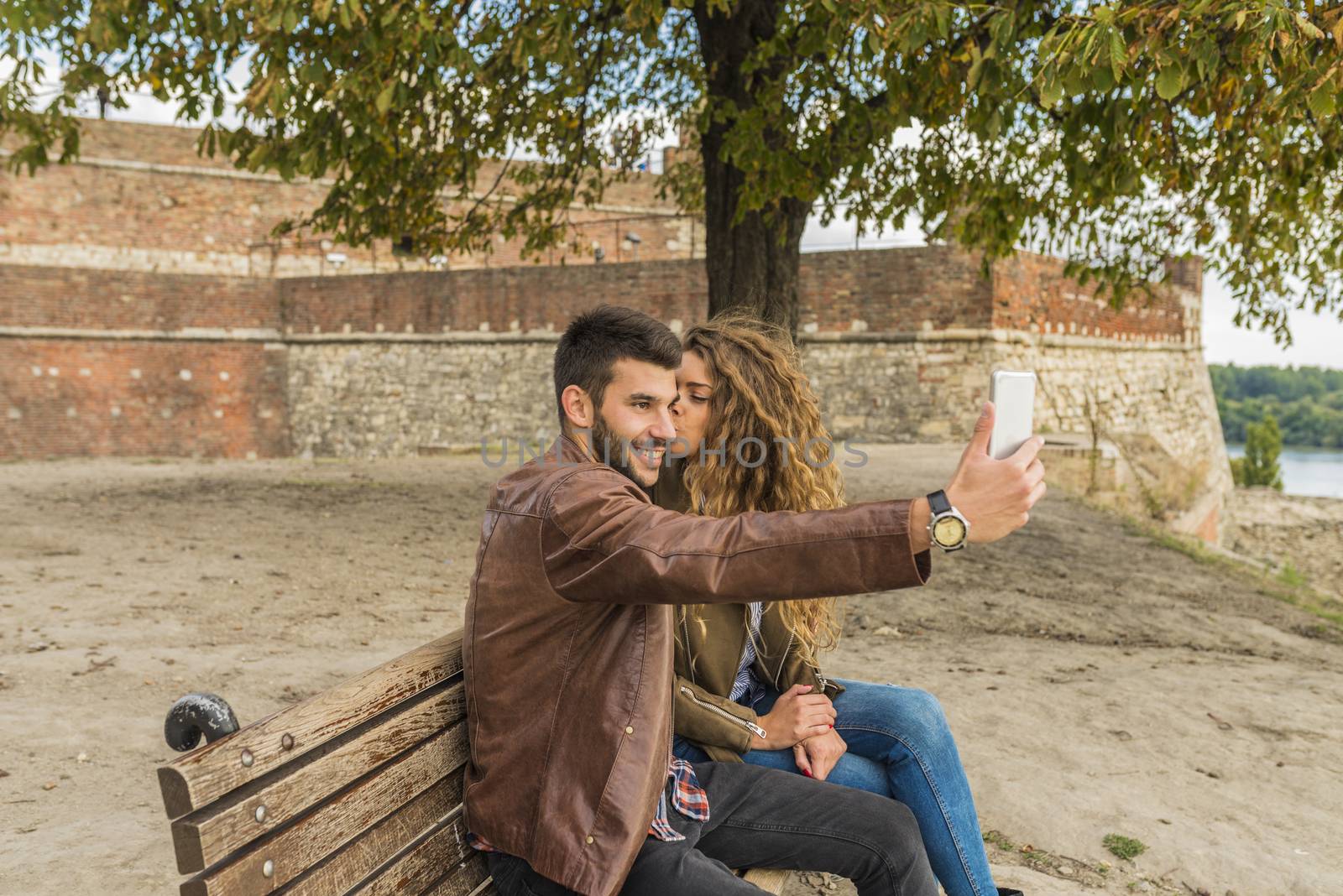 Young man is taking selfie while sitting on the bench in the park. His attractive girlfriend is kissing him.