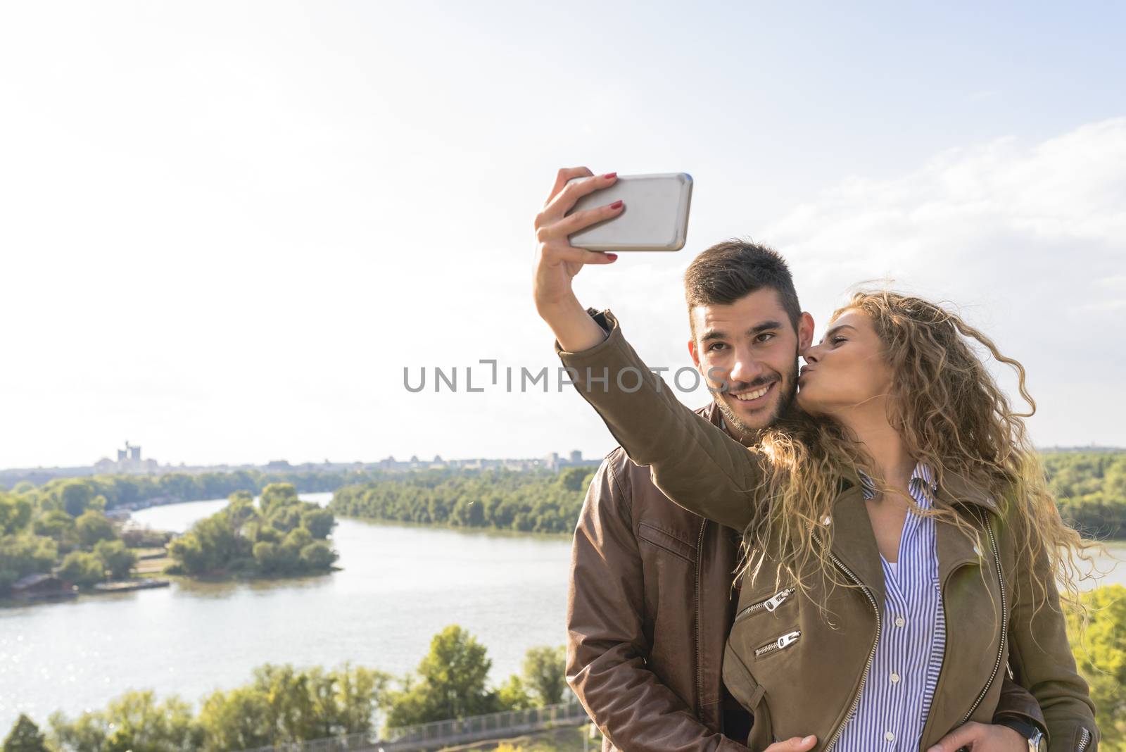 Beautiful young woman taking selfie in the nature while kissing her handsome boyfriend