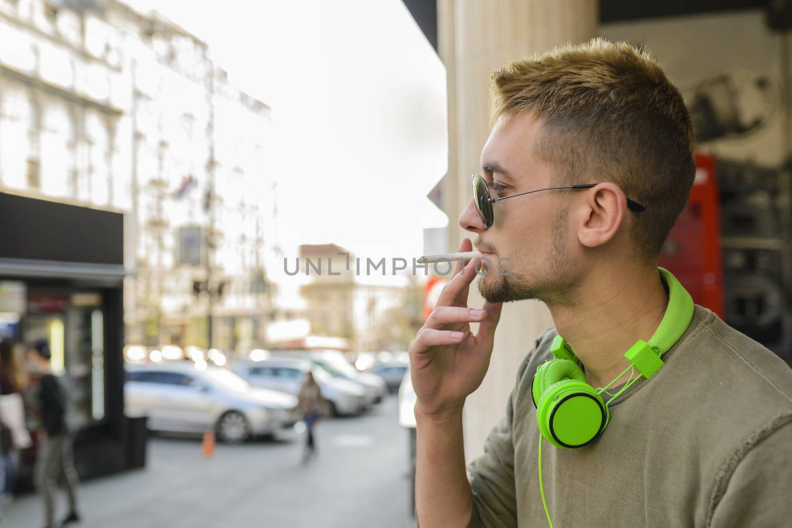 Young handsome man with green headphones and sunglasses enjoying a cigarette in the street.