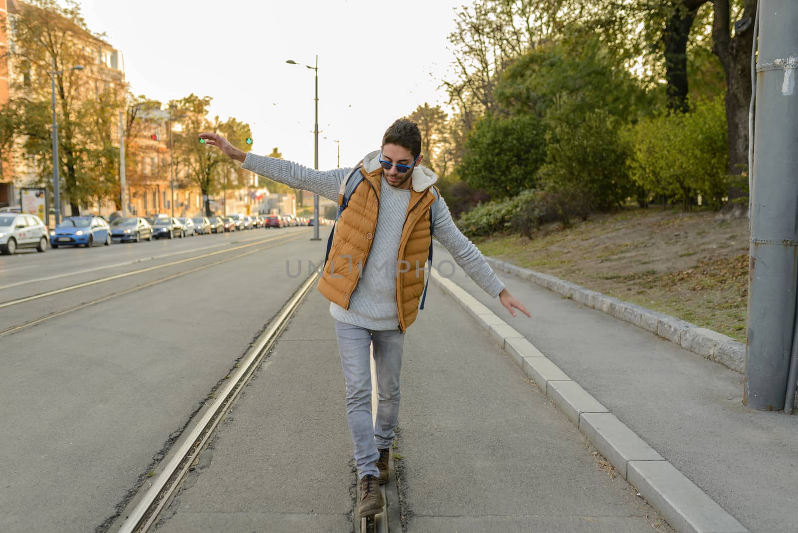 Young handsome hipster in orange jacket walking and balancing on the tramway rails in the city