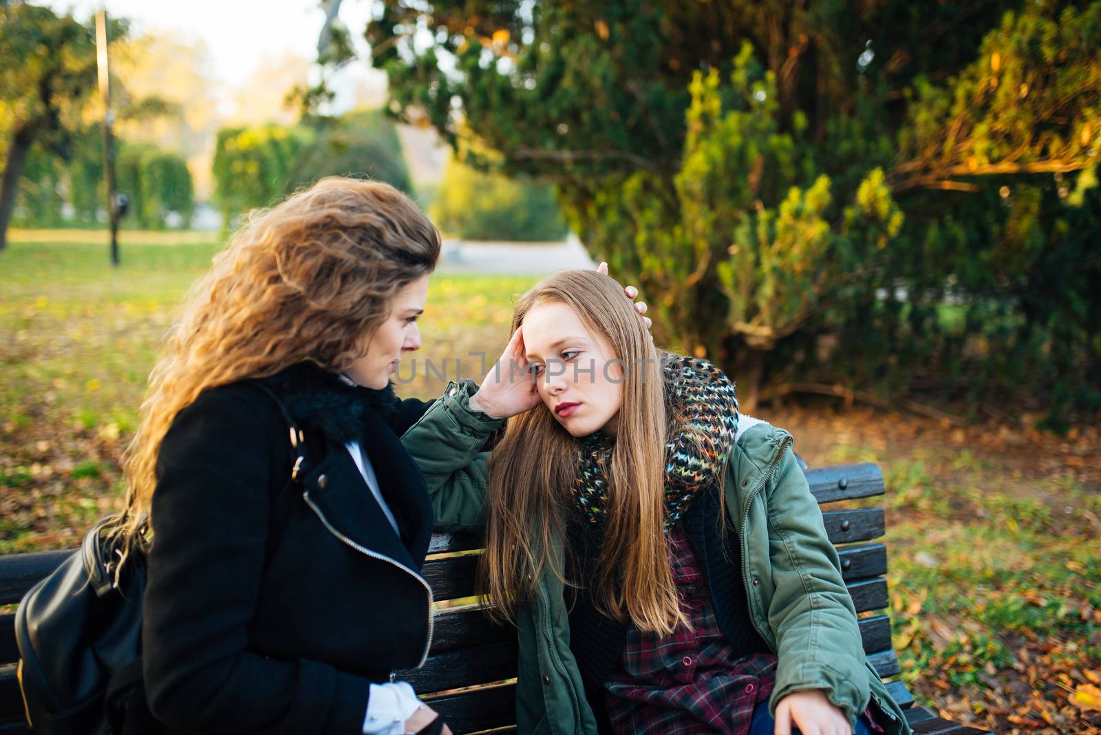Sad attractive girl sitting on the bench in the park while her female friend is consoling her.