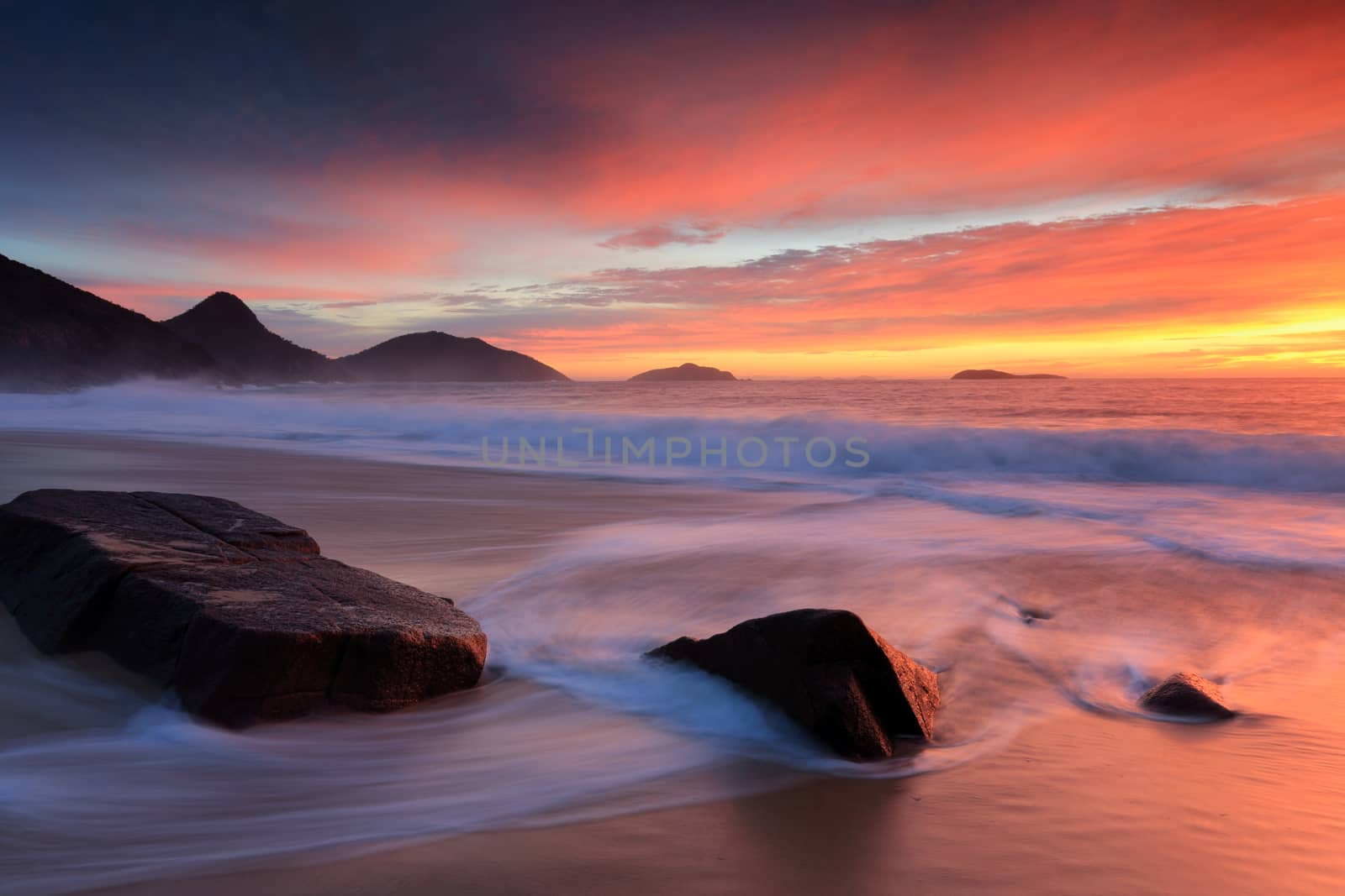 Mesmerising sunrise lighting up the sky in rich reds and in the water too as waves wash in around rocks on the sandy shoreline