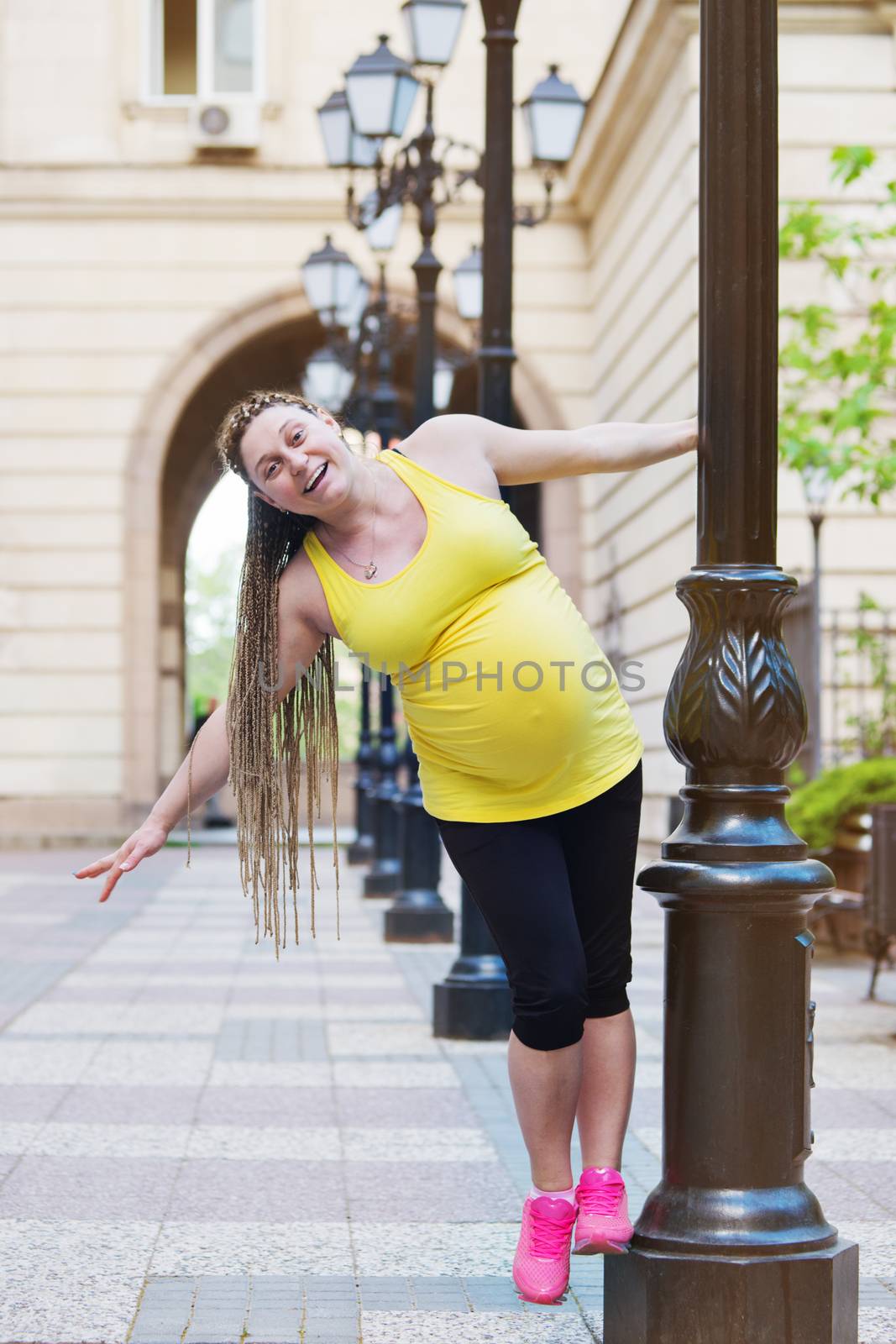 Smiling happy pregnant woman is playing on a street pole.