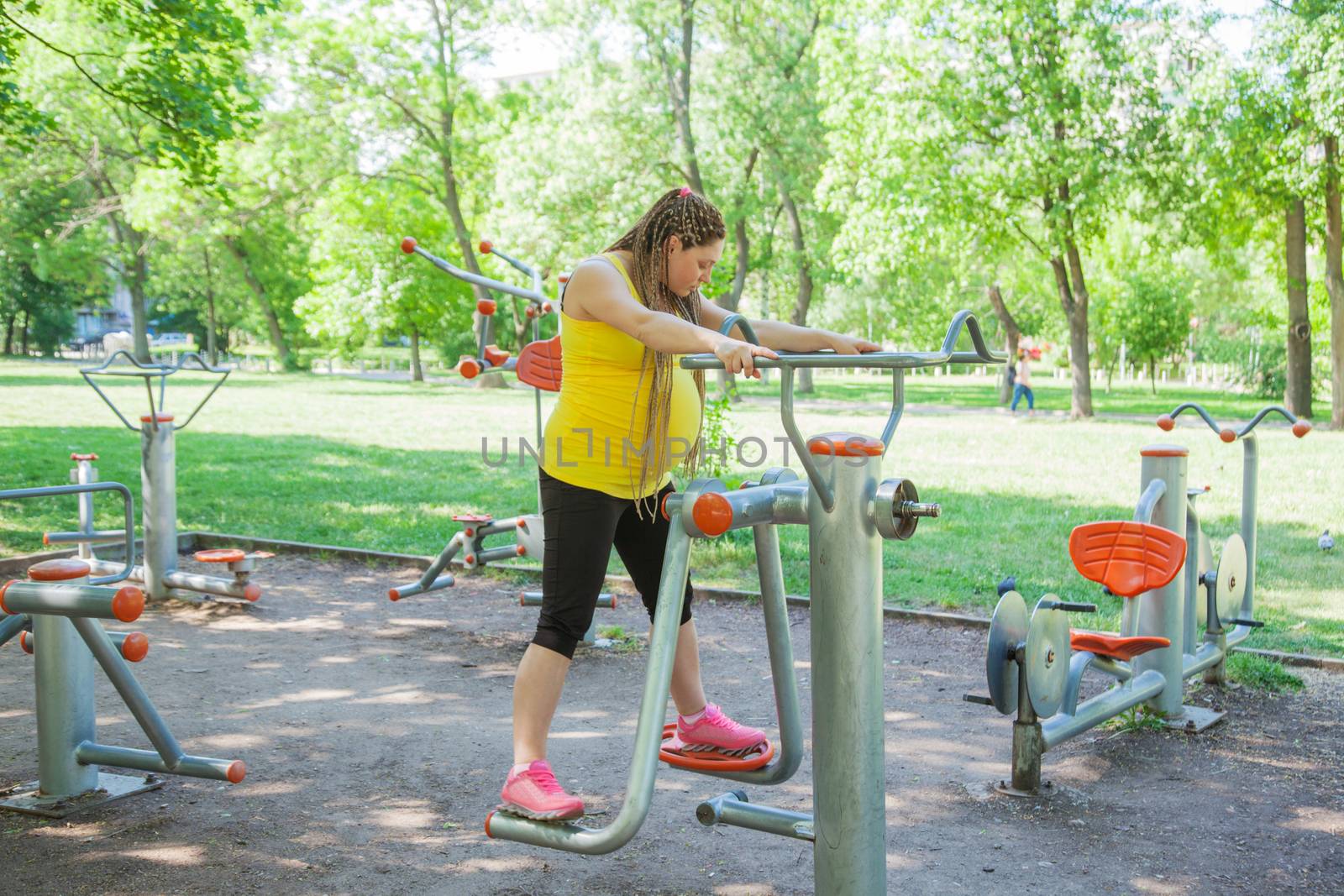 Pregnant woman is doing exercises outdoors on fitness machine in a park.