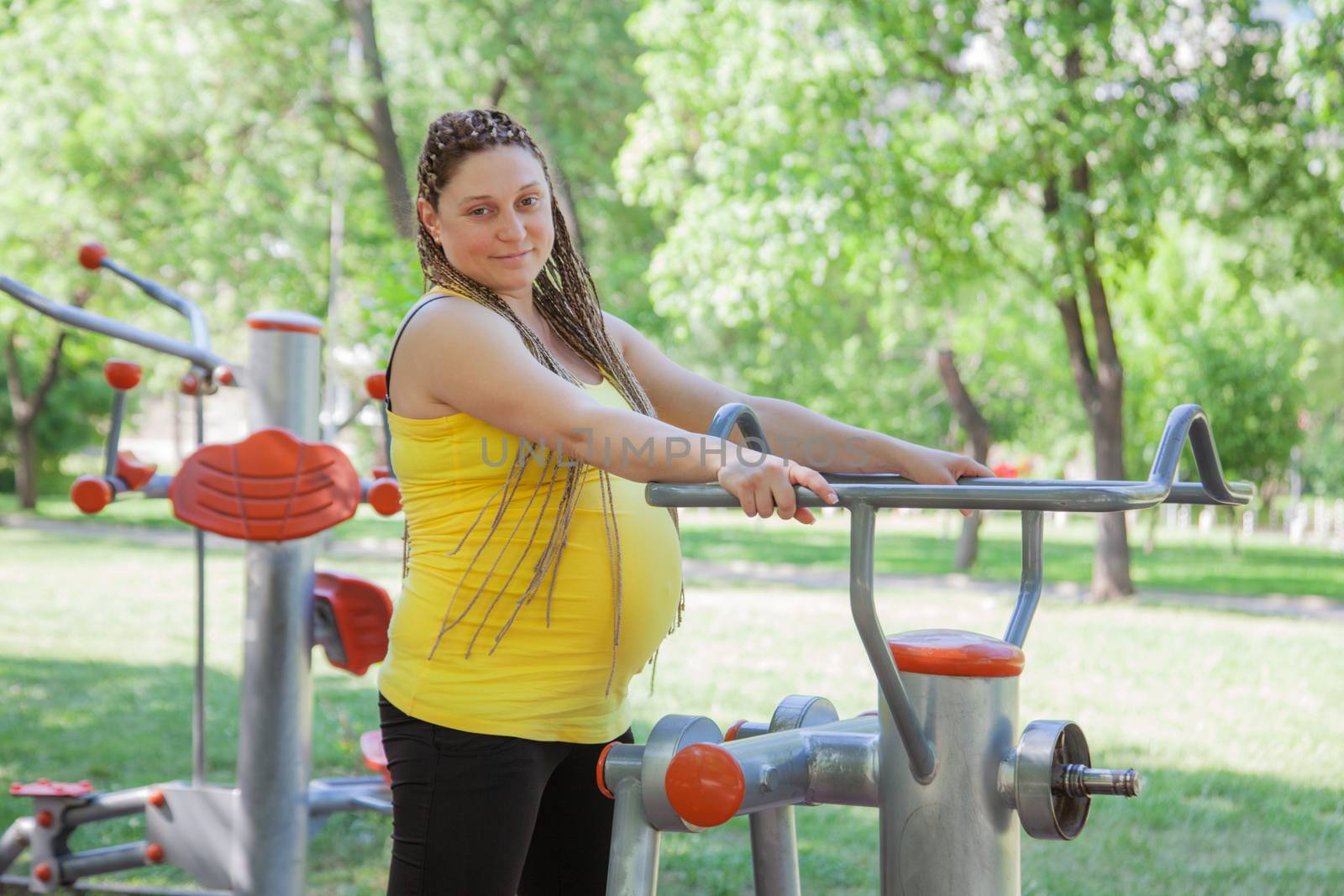 Pregnant woman is exercising outdoors on public fitness machines.