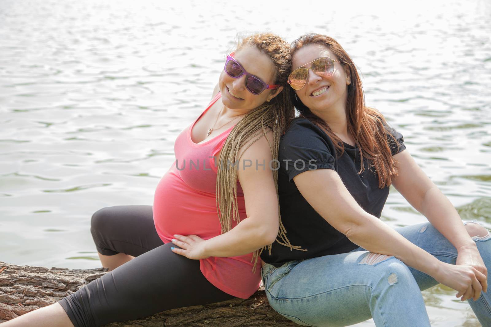 Girlfriends are sitting on a wooden log in front of water, one female is pregnant.