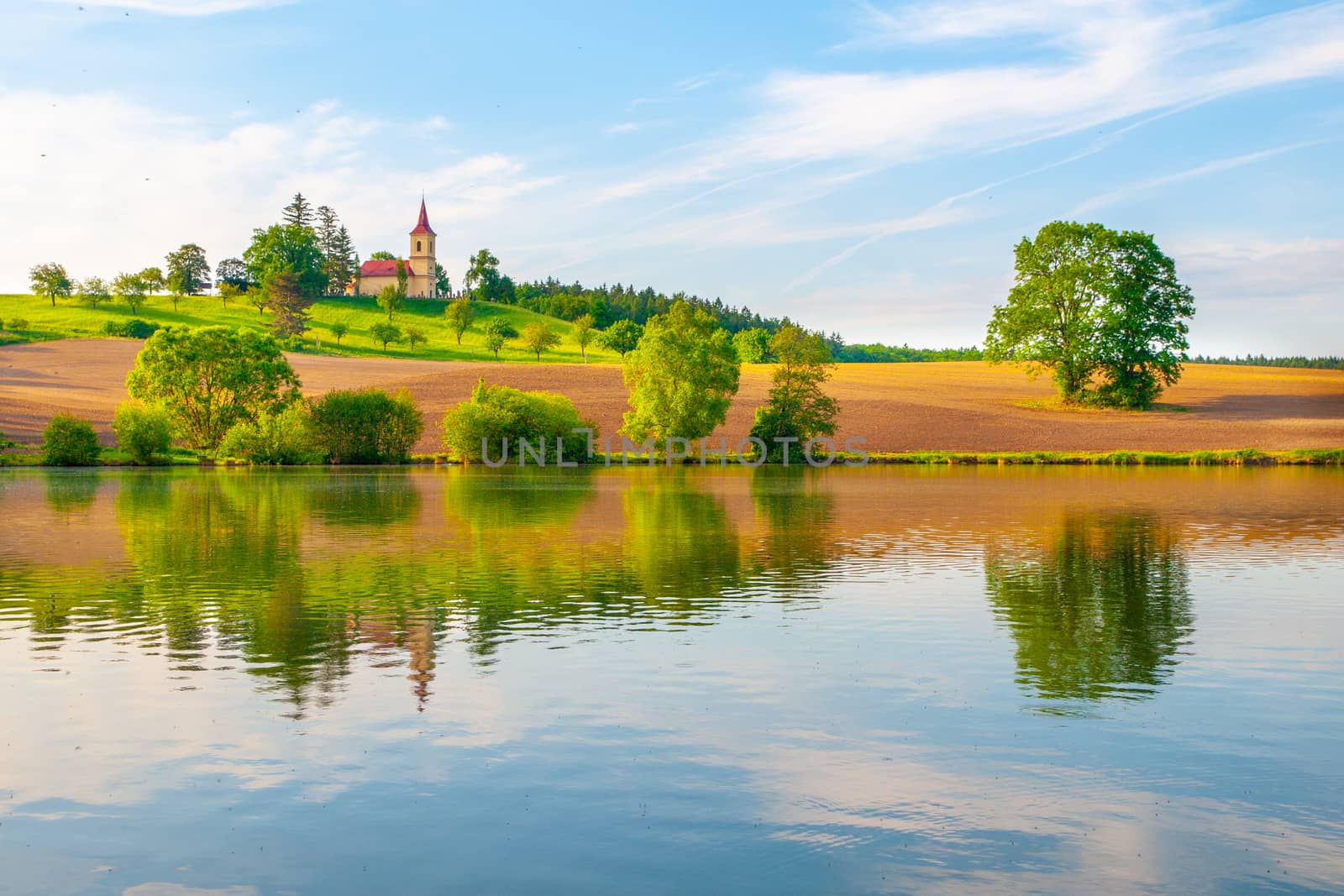 Romantic landscape with small church on the hill reflected in the pond. Sunny summer day with blue sky and white clouds. St. Peter and Pauls church at Bysicky near Lazne Belohrad, Czech Republic.