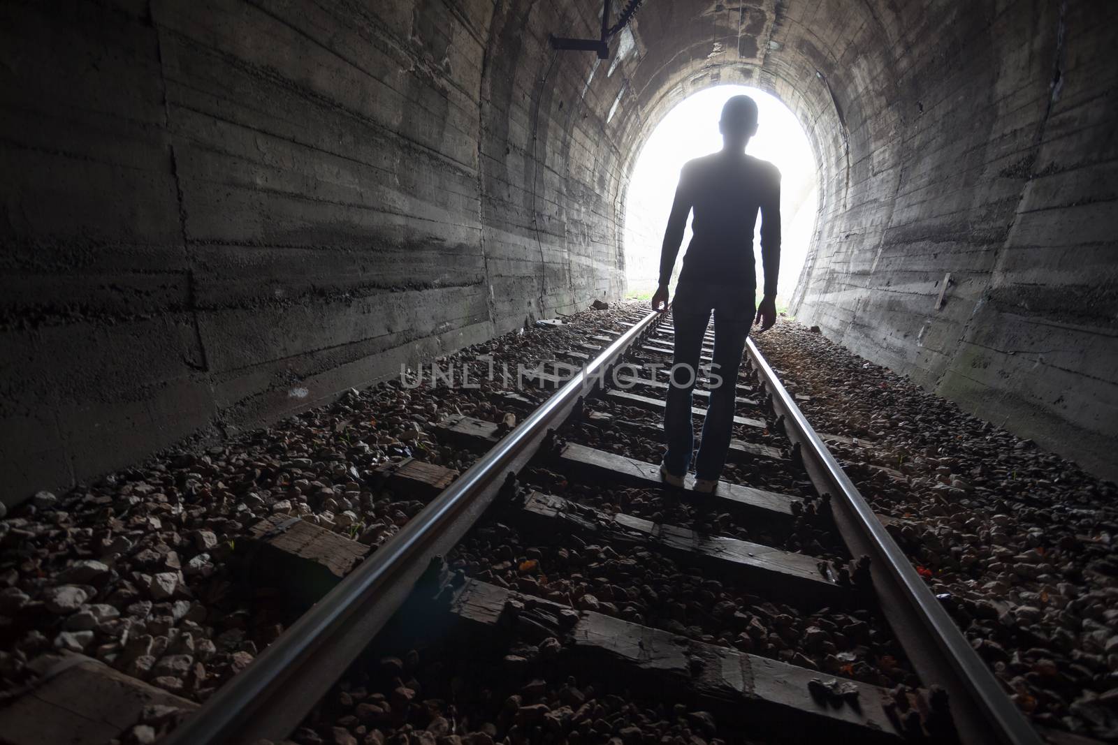 Man silhouetted in a tunnel standing in the center of the railway tracks looking towards the light at the end of the tunnel in a conceptual image