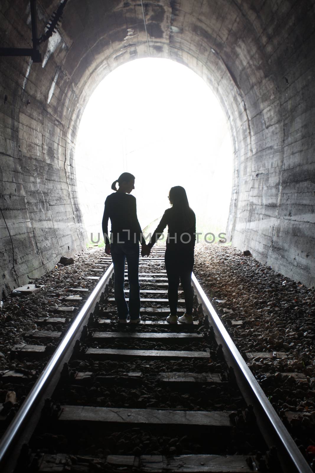 Couple walking hand in hand along the track through a railway tunnel towards the bright light at the other end, they appear as silhouettes against the light