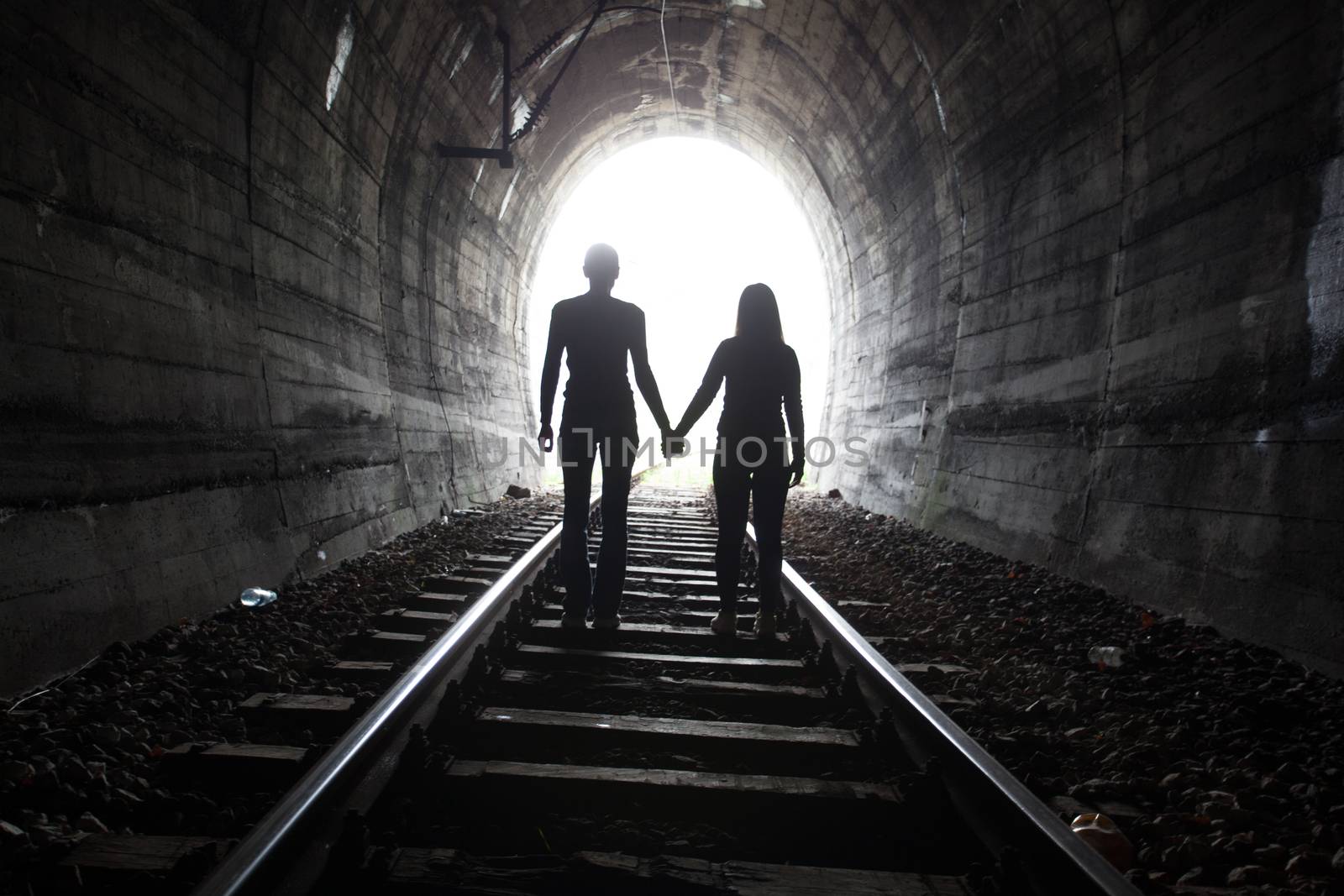 Couple walking hand in hand along the track through a railway tunnel towards the bright light at the other end, they appear as silhouettes against the light