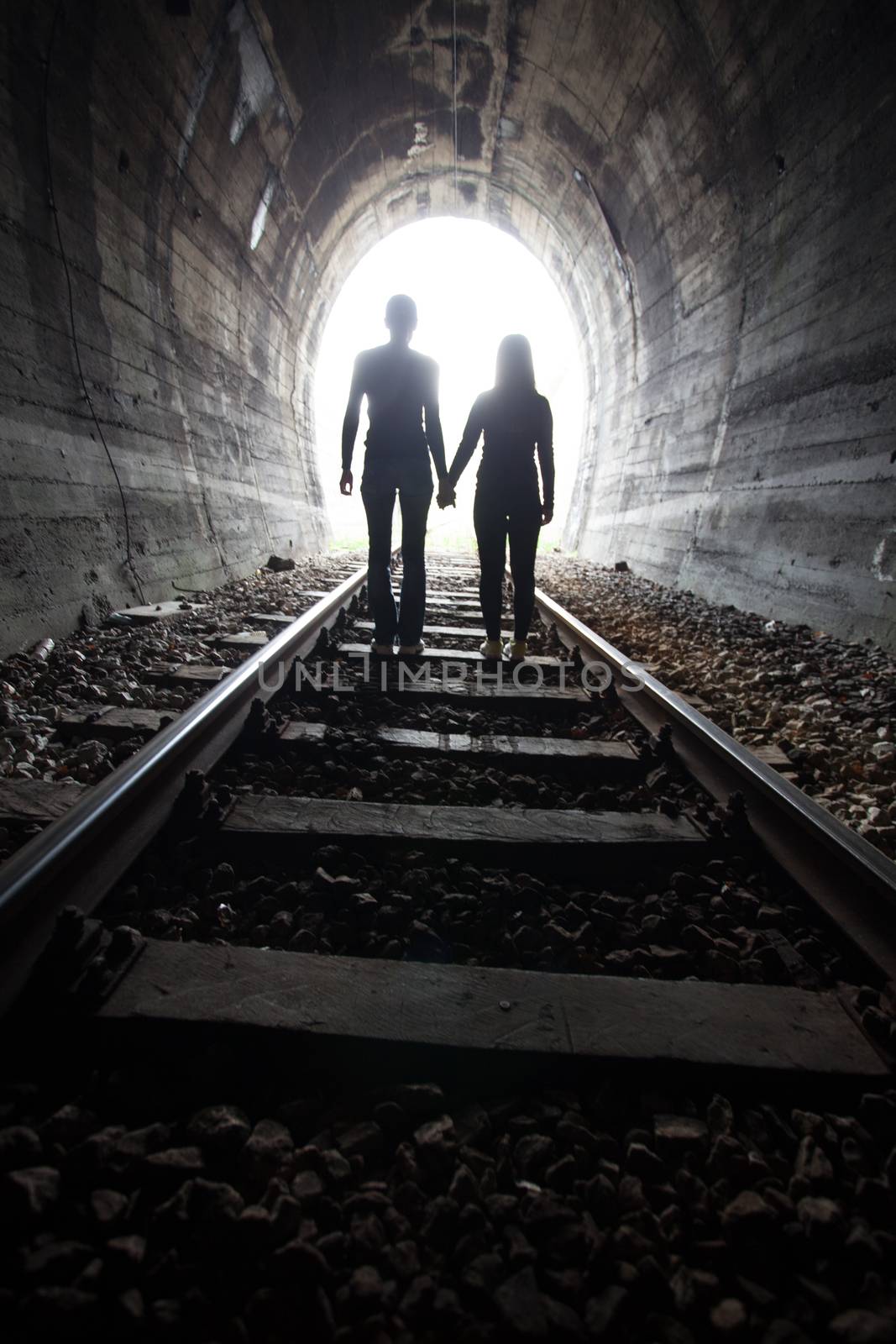 Couple walking hand in hand along the track through a railway tunnel towards the bright light at the other end, they appear as silhouettes against the light