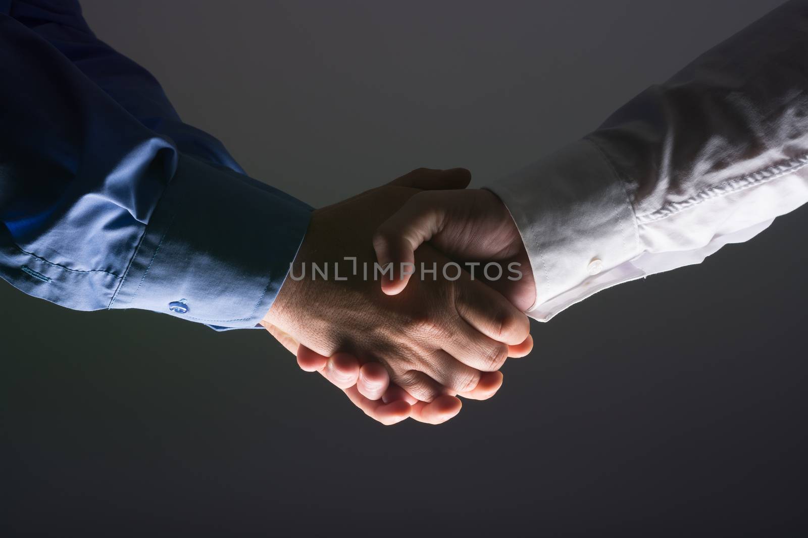 Close up side view of two male hands shaking over dark gray background and light source at bottom side