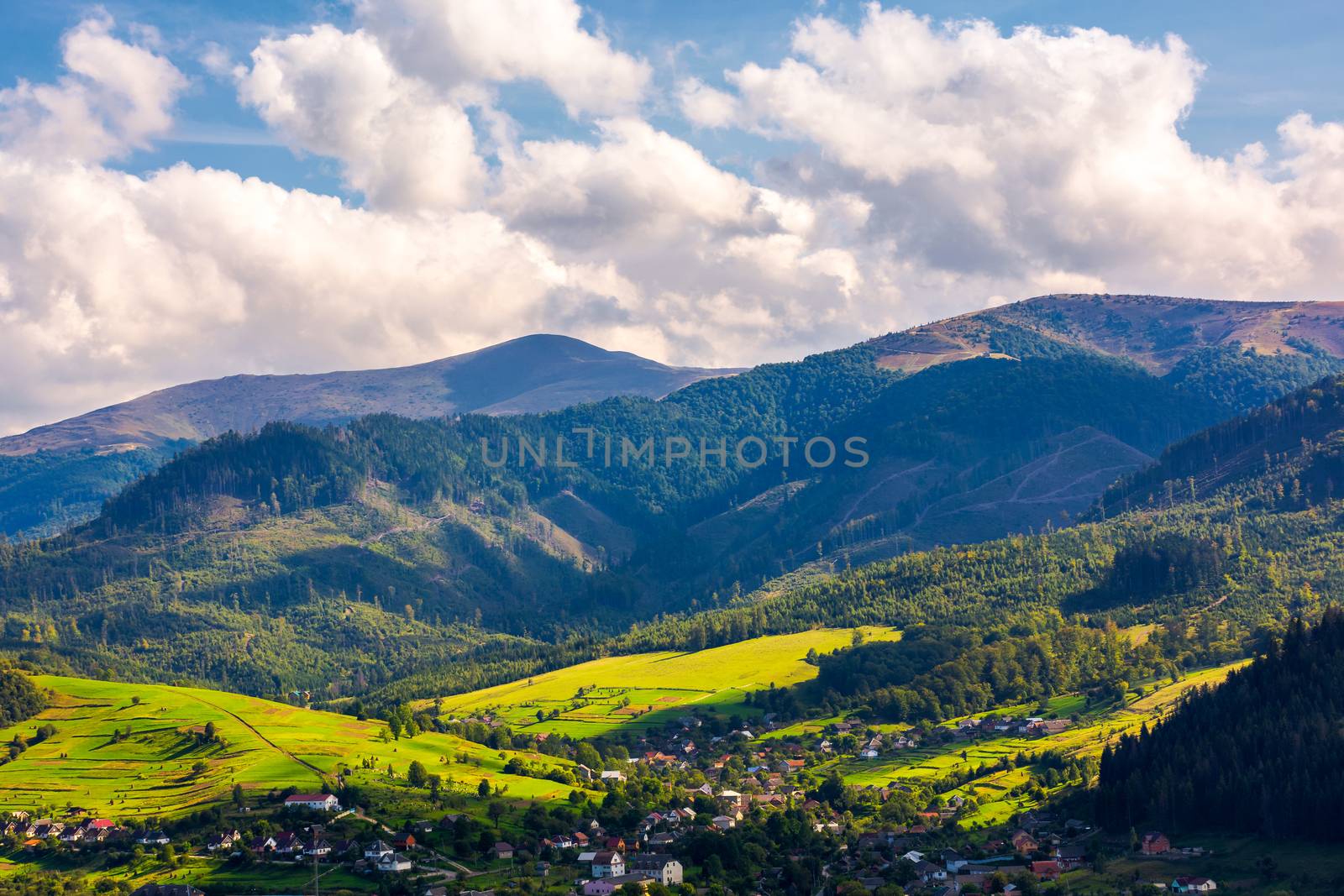 beautiful urban landscape in mountains. village at the foot of the mountain. interesting cloud formation over the ridge. grassy rural fields on hills. lovely sunny afternoon